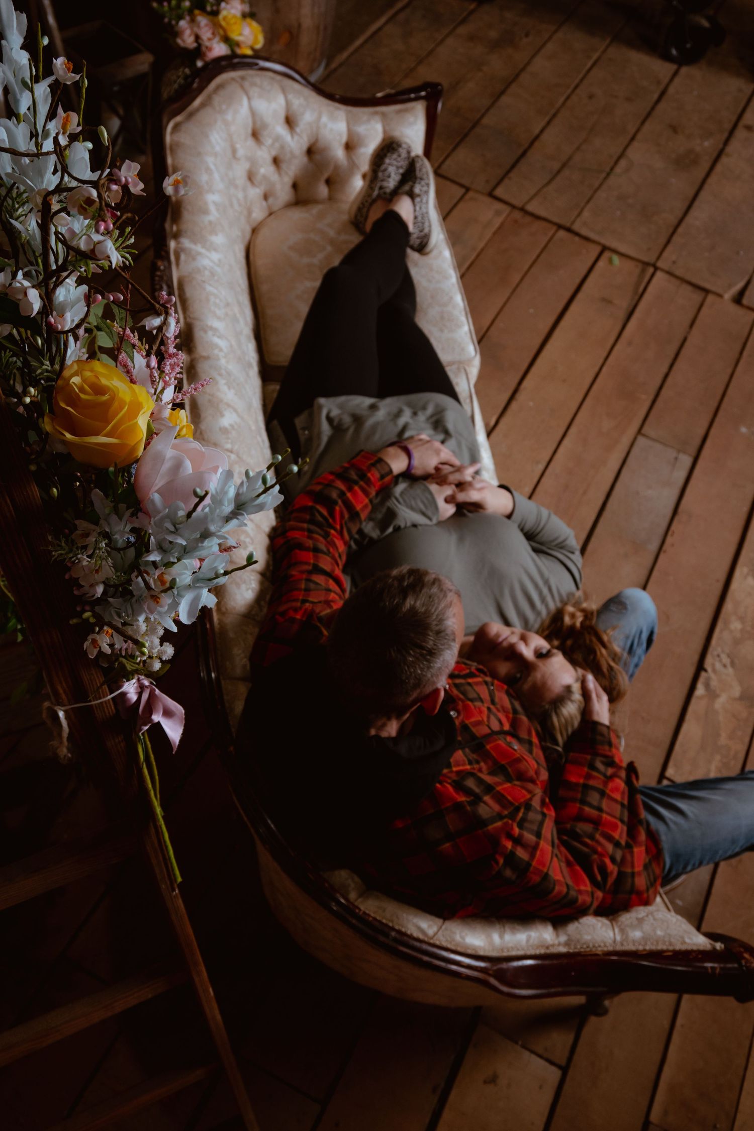 Couple lying together on a vintage chair surrounded by colorful flowers on a rustic wooden floor.