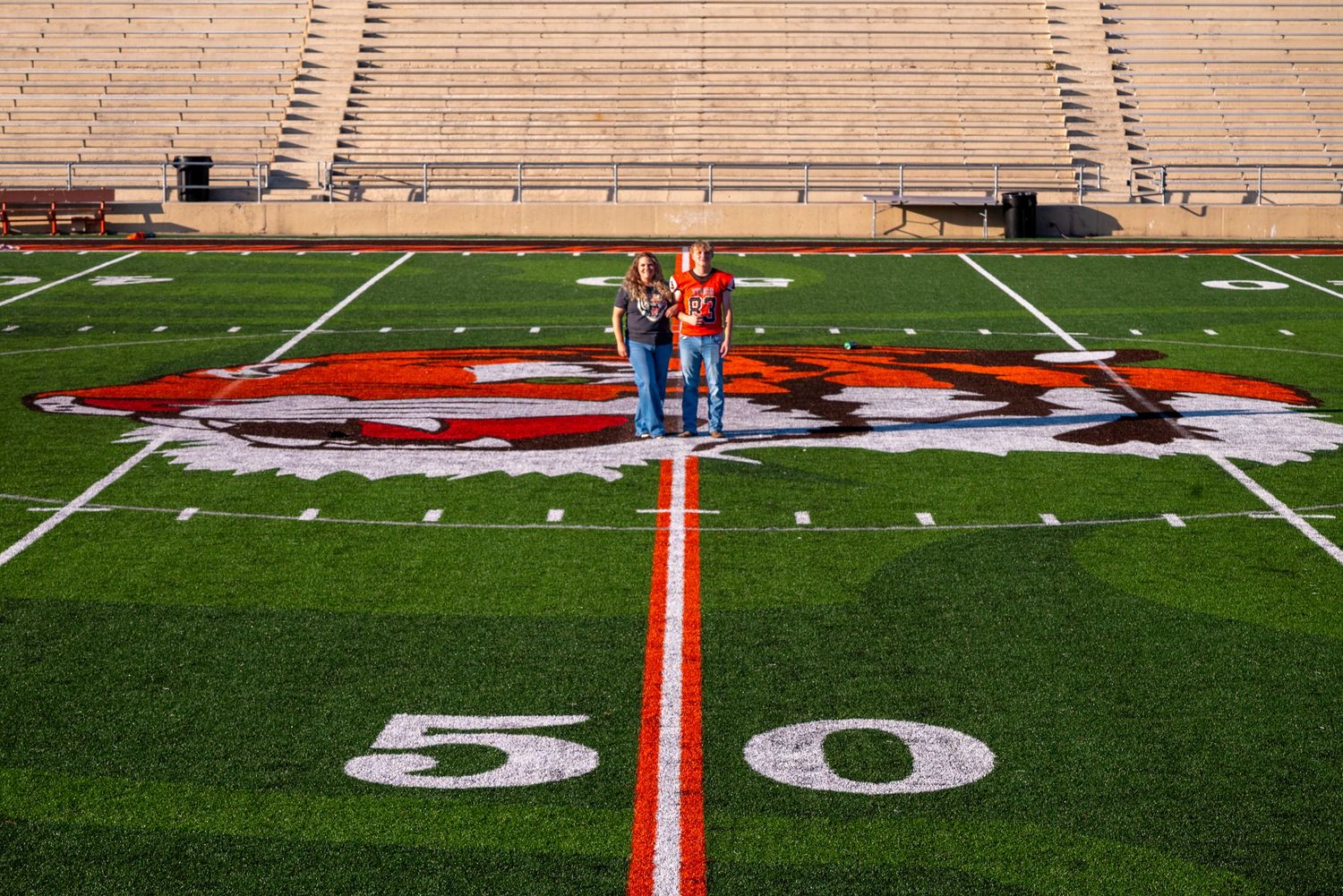 Two people stand on the 50-yard line of a red and white football stadium field with large mascot logo.