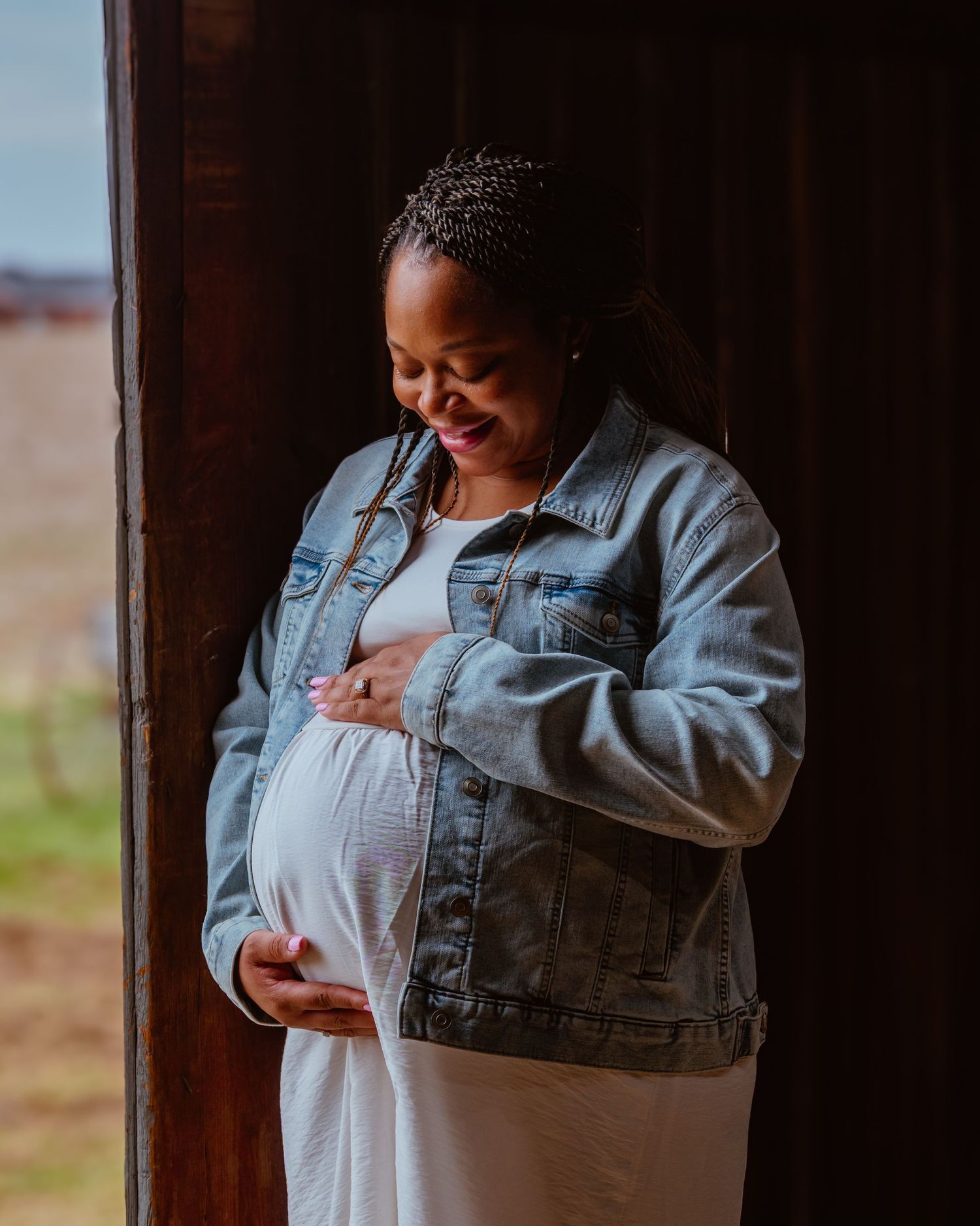 Smiling pregnant Black woman in denim jacket and white dress cradling her baby bump while leaning against a wooden wall.