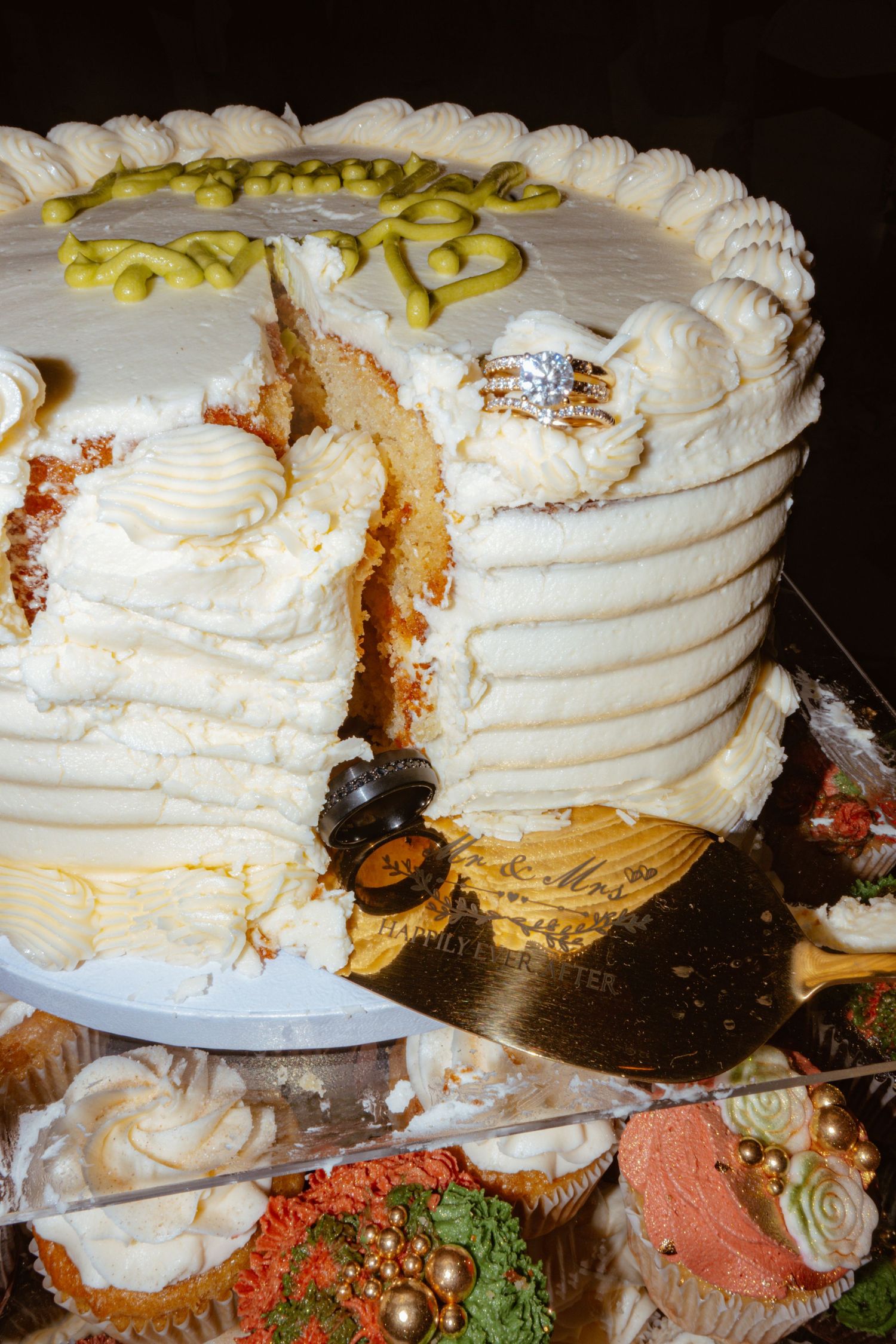 Elegant white wedding cake with gold accents being sliced, surrounded by decorated cupcakes.