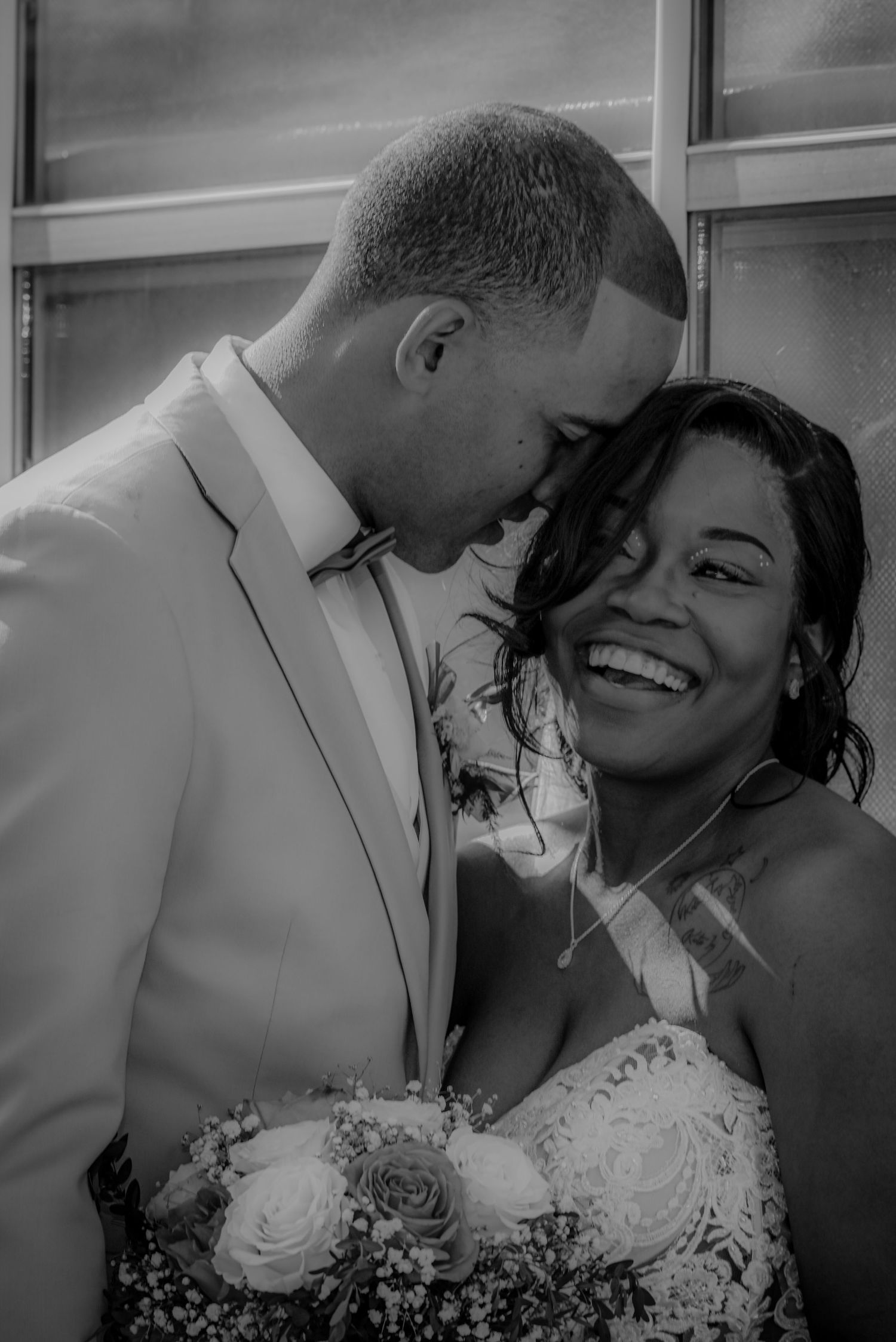 Black and white wedding photo of smiling bride holding bouquet leaning close with groom in white suit.
