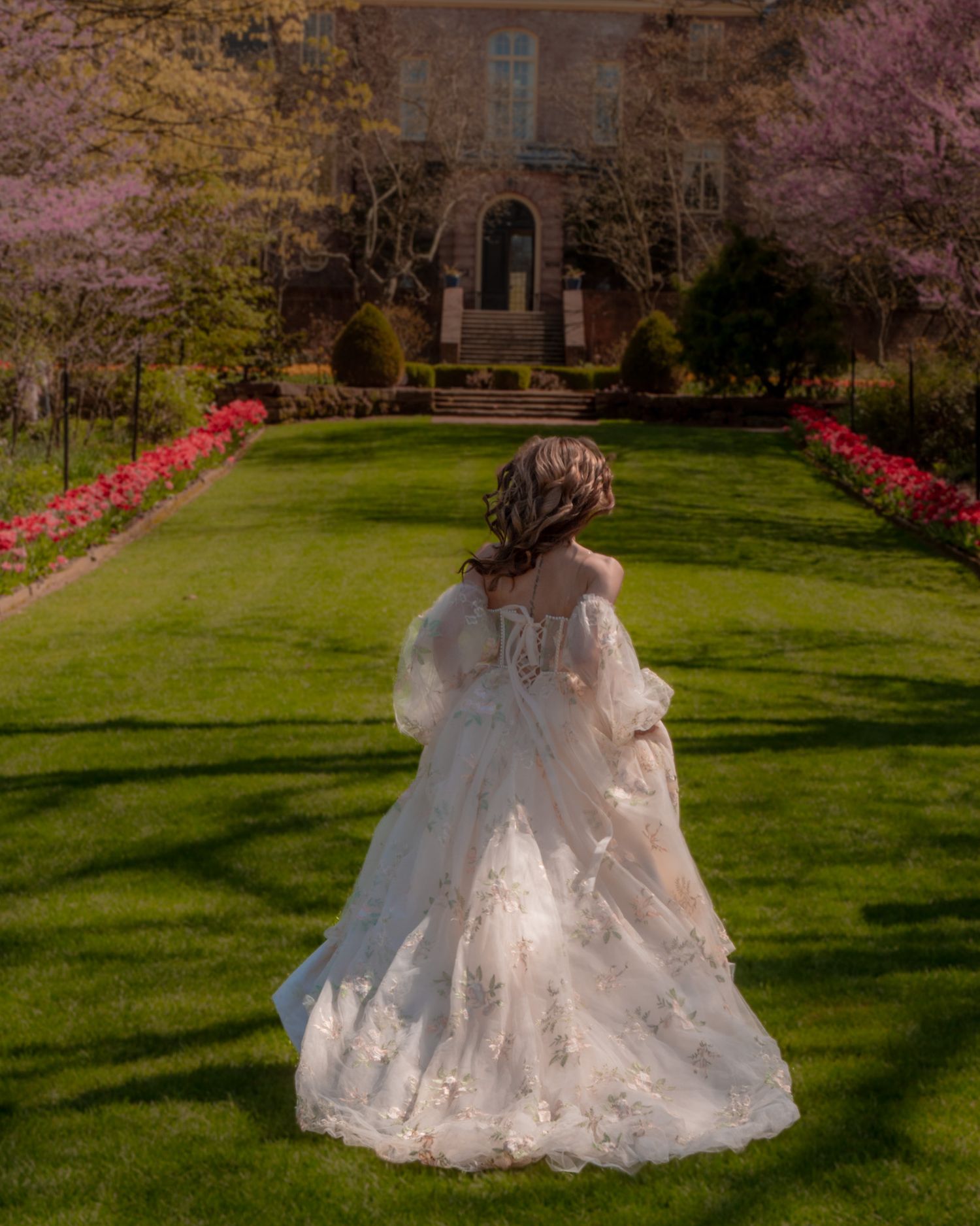 Back view of a bride in a flowing lace wedding gown standing on manicured lawn with blooming gardens and mansion backdrop.