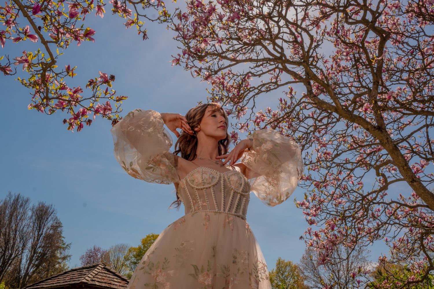 Woman in elegant white corset dress with ruffled sleeves poses under blooming pink magnolia trees on a sunny day.