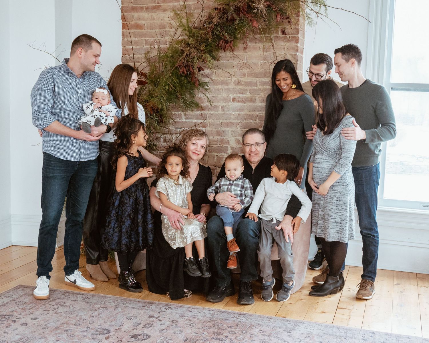 Large multigenerational family posing together in a bright studio with brick wall backdrop and greenery decor.