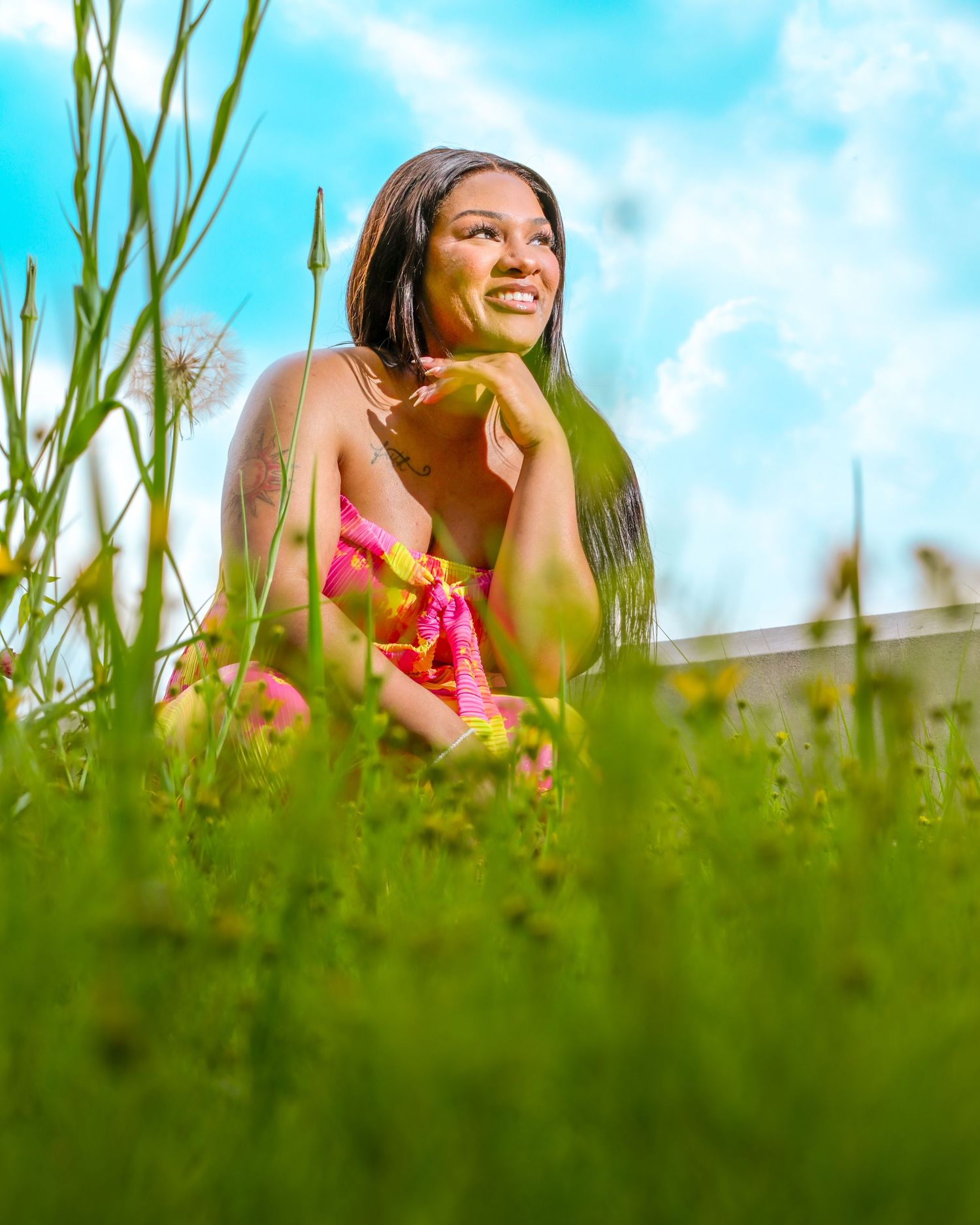 A joyful person in a pink top reclines in a sun-dappled meadow with tall grass and wildflowers under blue summer skies.