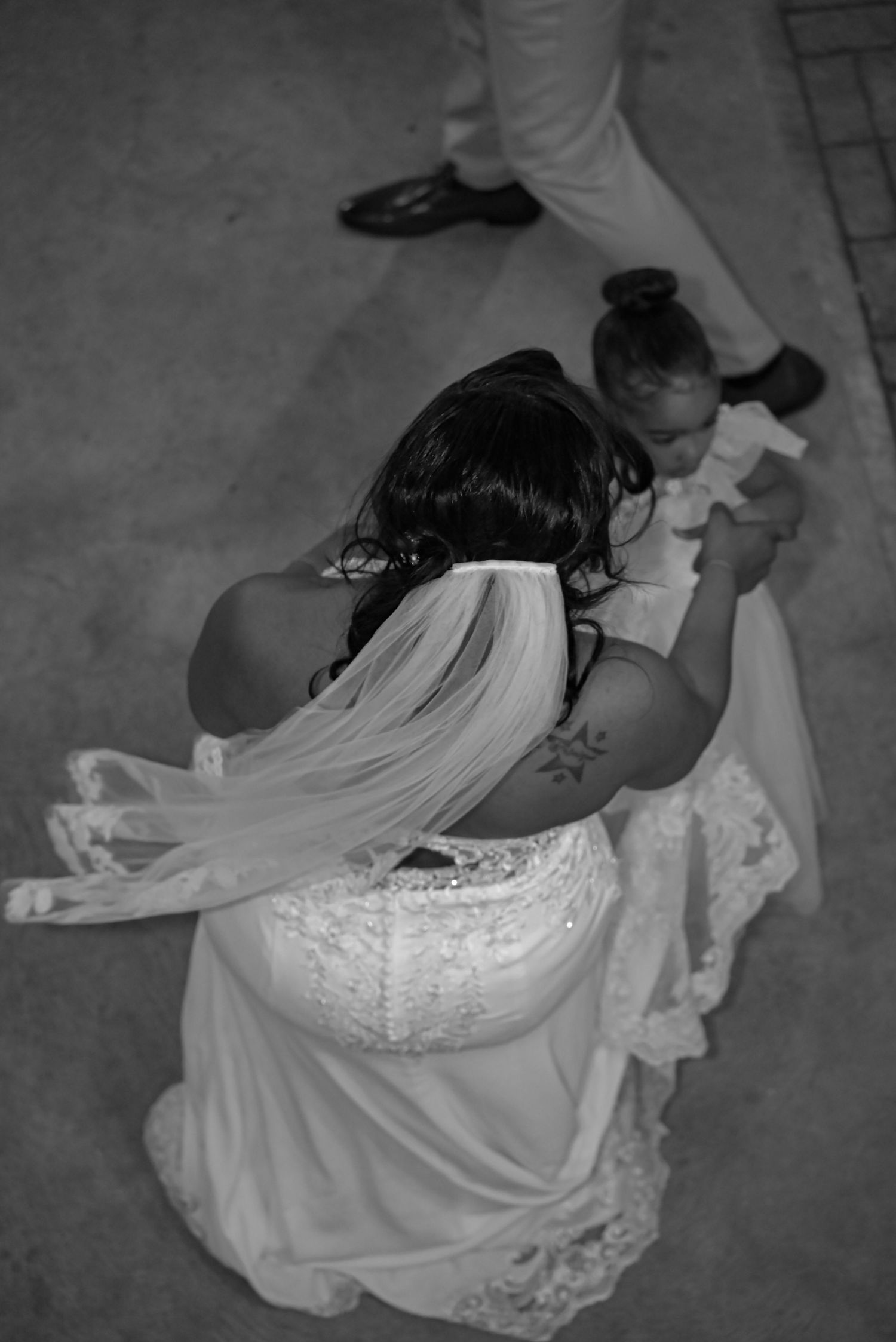 Black and white aerial photo of a bride in lace wedding dress and veil dancing with a young child at a wedding reception.