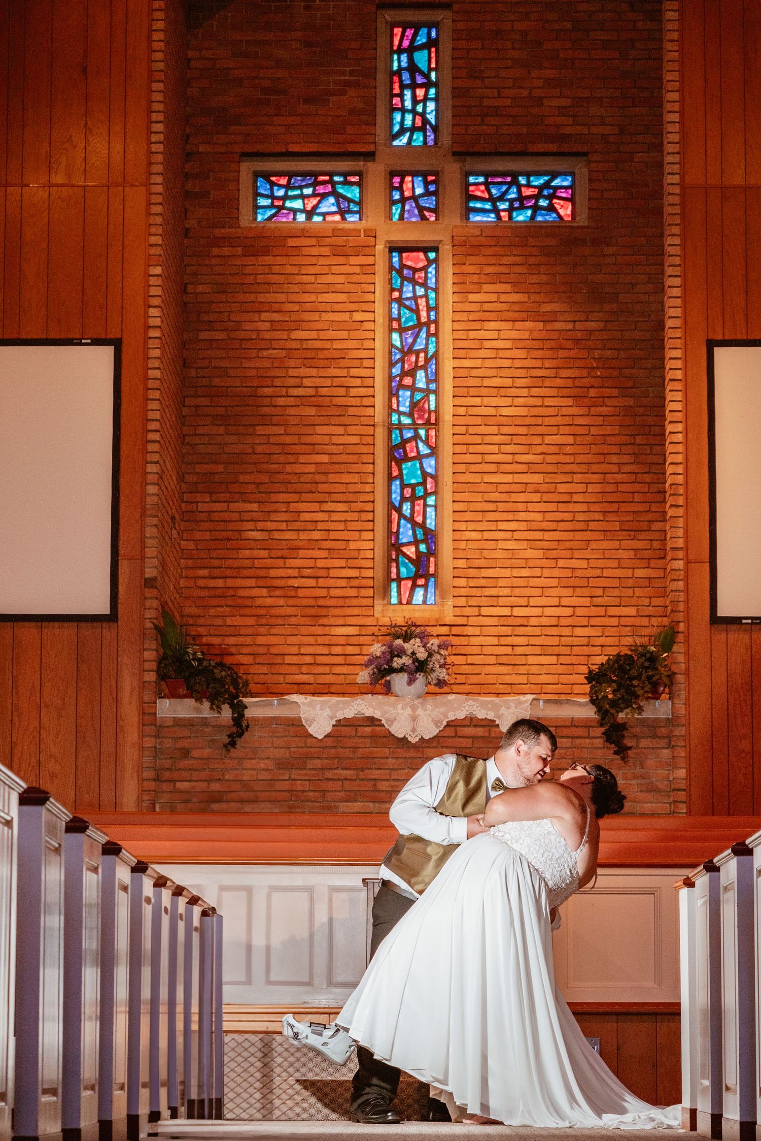 Bride and groom share a romantic dip kiss at church altar with colorful stained glass cross backdrop.