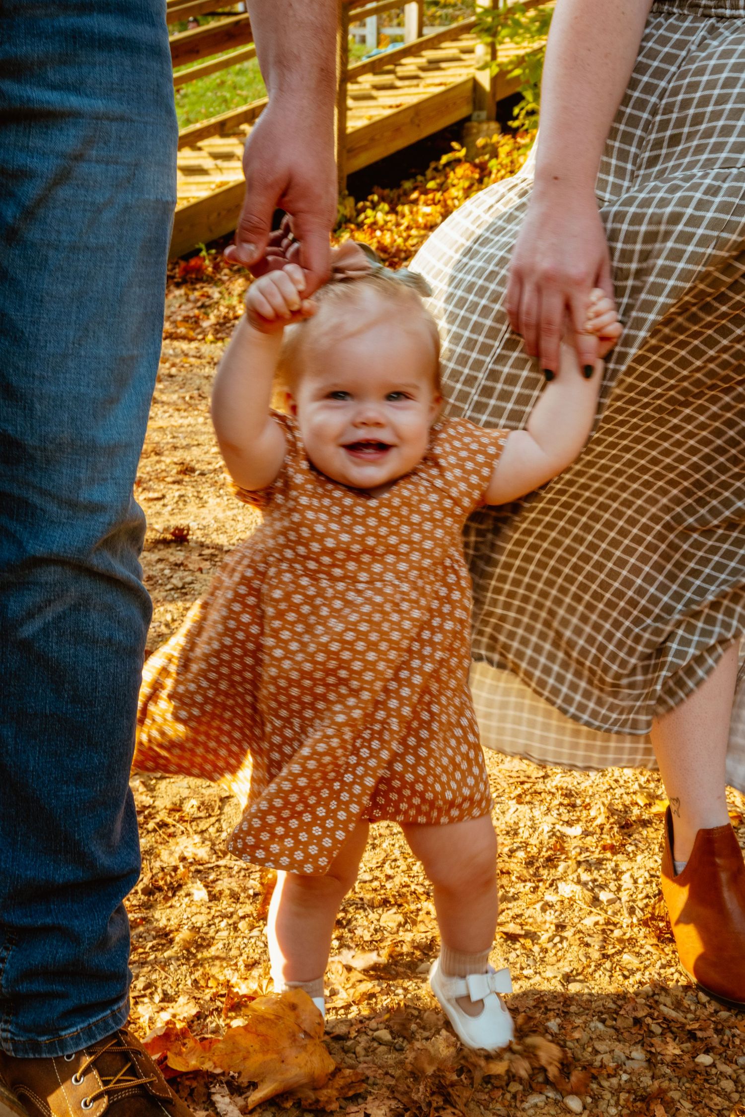 Smiling baby girl in polka dot dress takes first steps outdoors in autumn, held by parents hands.