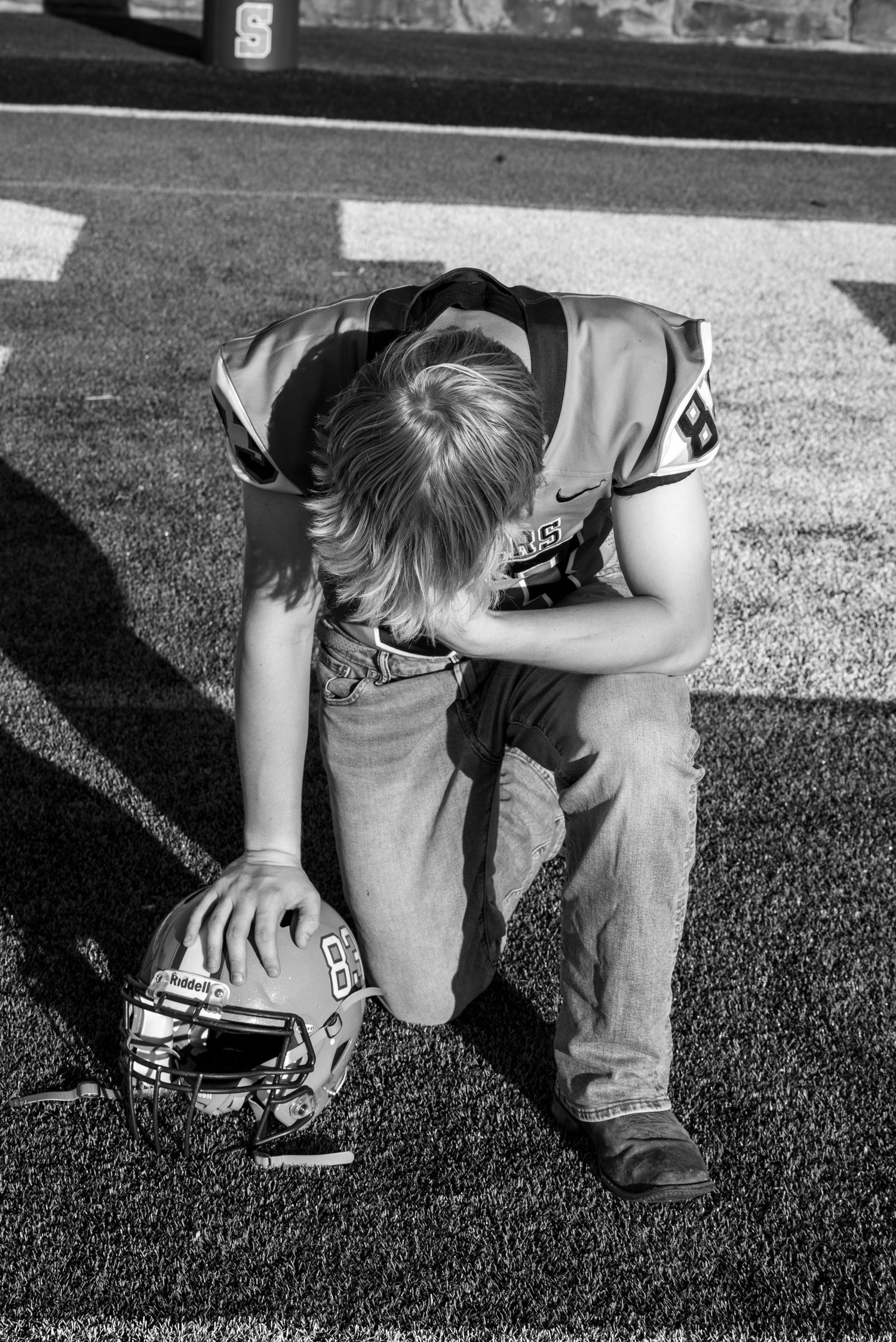 Black and white photo of a young football player kneeling on field, holding helmet, head bowed in reflection.