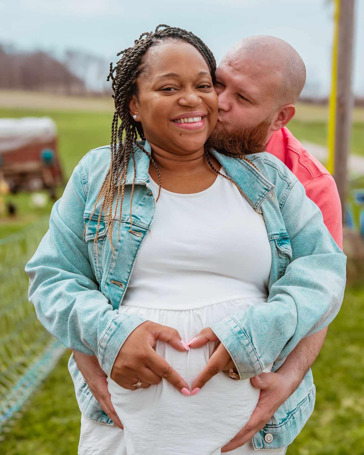 Couple sharing a tender maternity photo outdoors, forming a heart shape over pregnant belly while man kisses womans cheek.