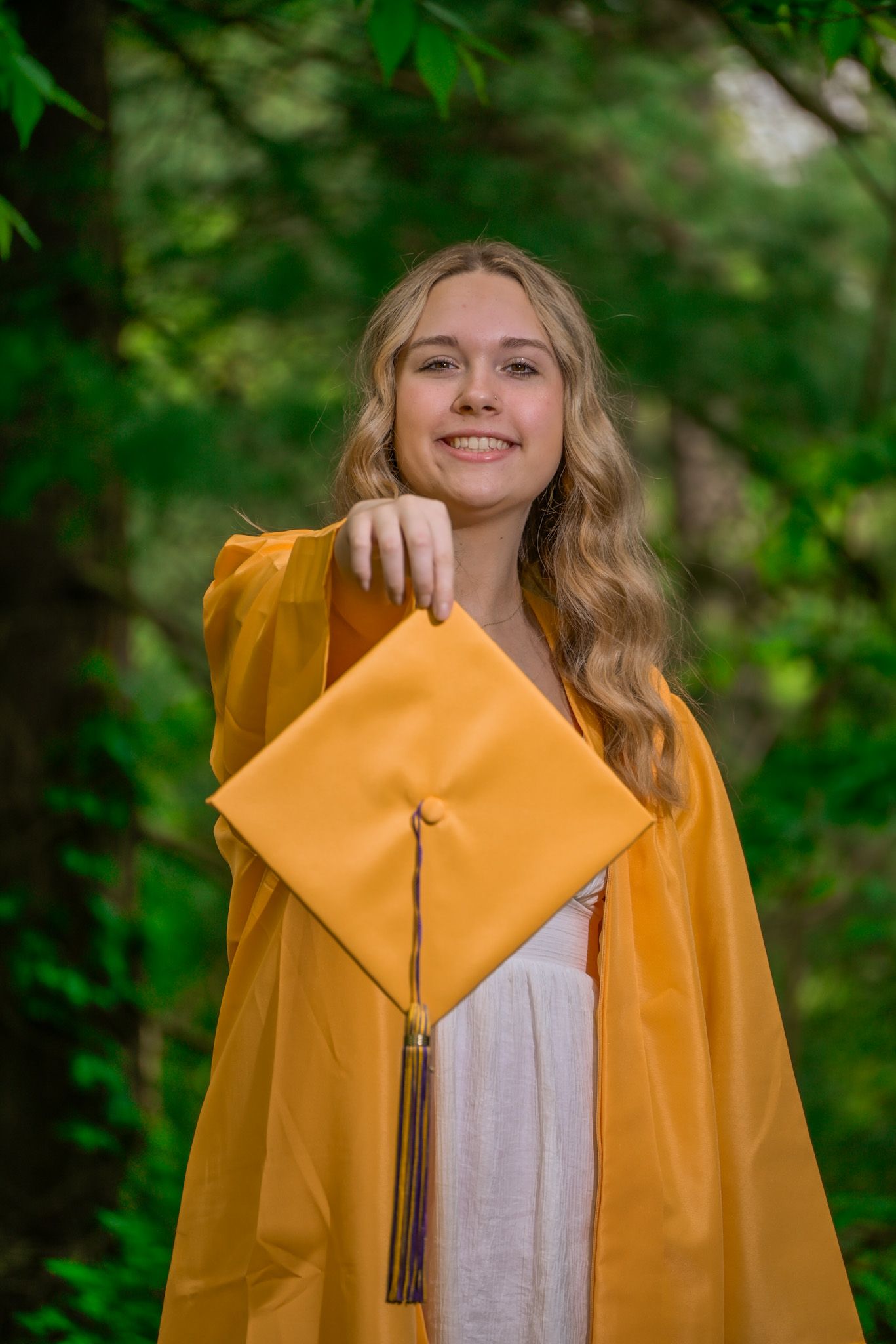 Smiling teenage girl in gold graduation cap and gown holding mortarboard toward camera outdoors.