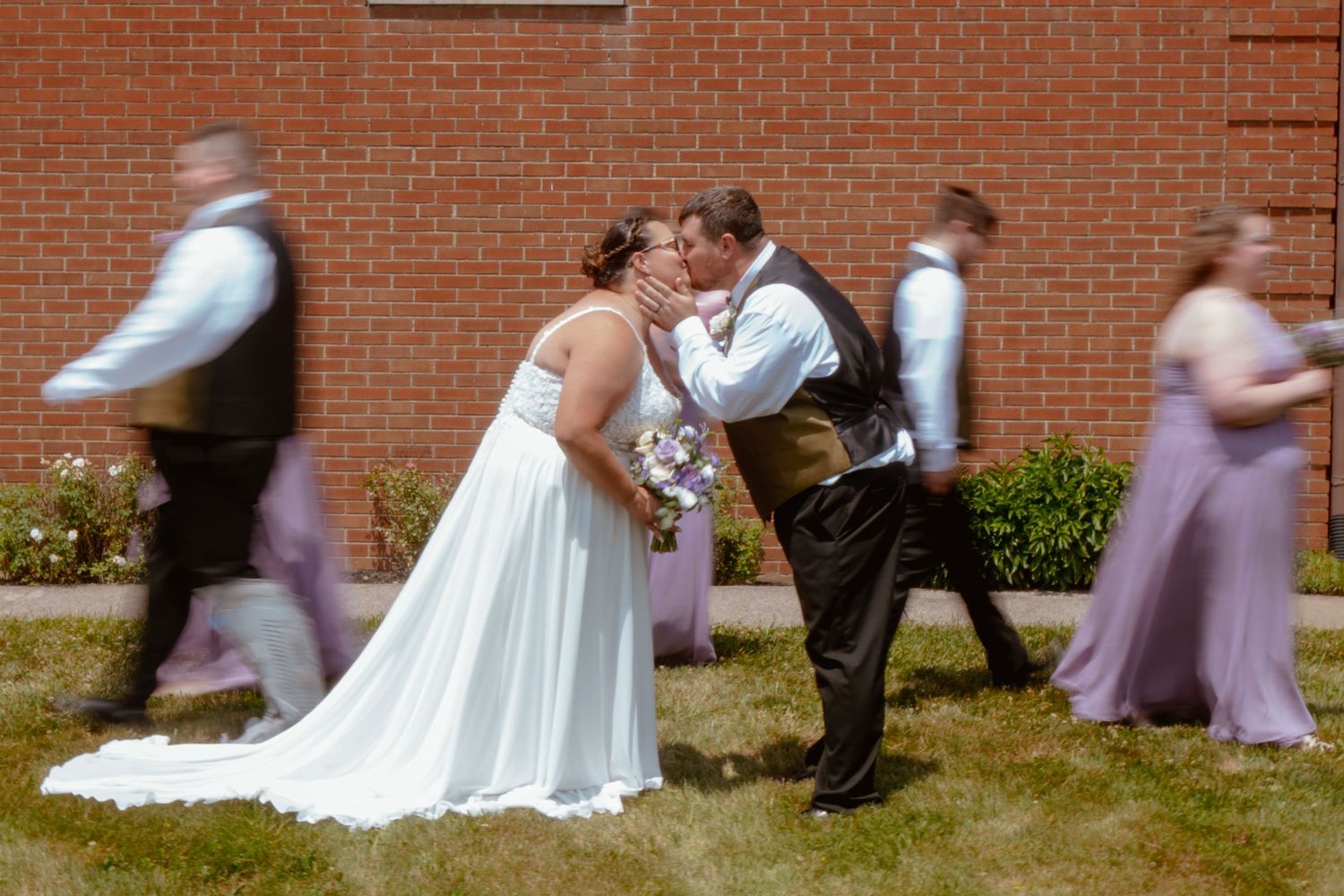 Bride and groom share a kiss while wedding party walks past brick wall backdrop on sunny day.