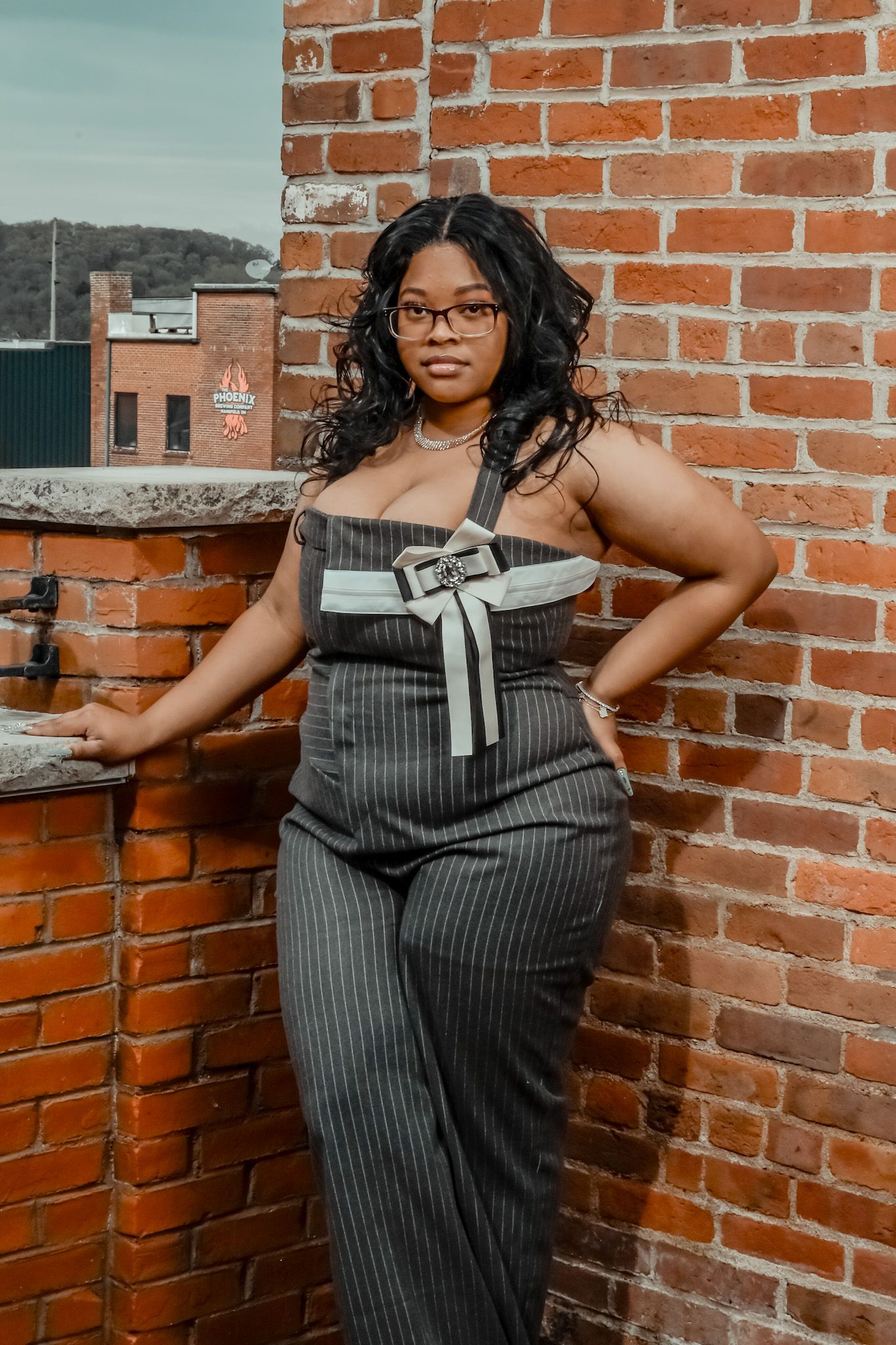 A woman in a pinstriped jumpsuit poses confidently against a rustic brick wall backdrop.