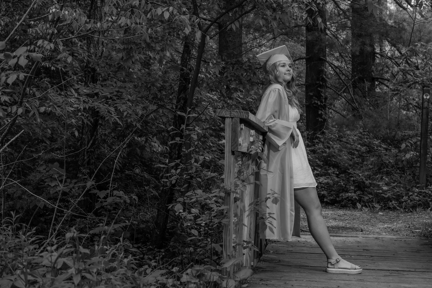 A woman in a white dress stands on a wooden bridge surrounded by lush forest trees in a black and white photo.