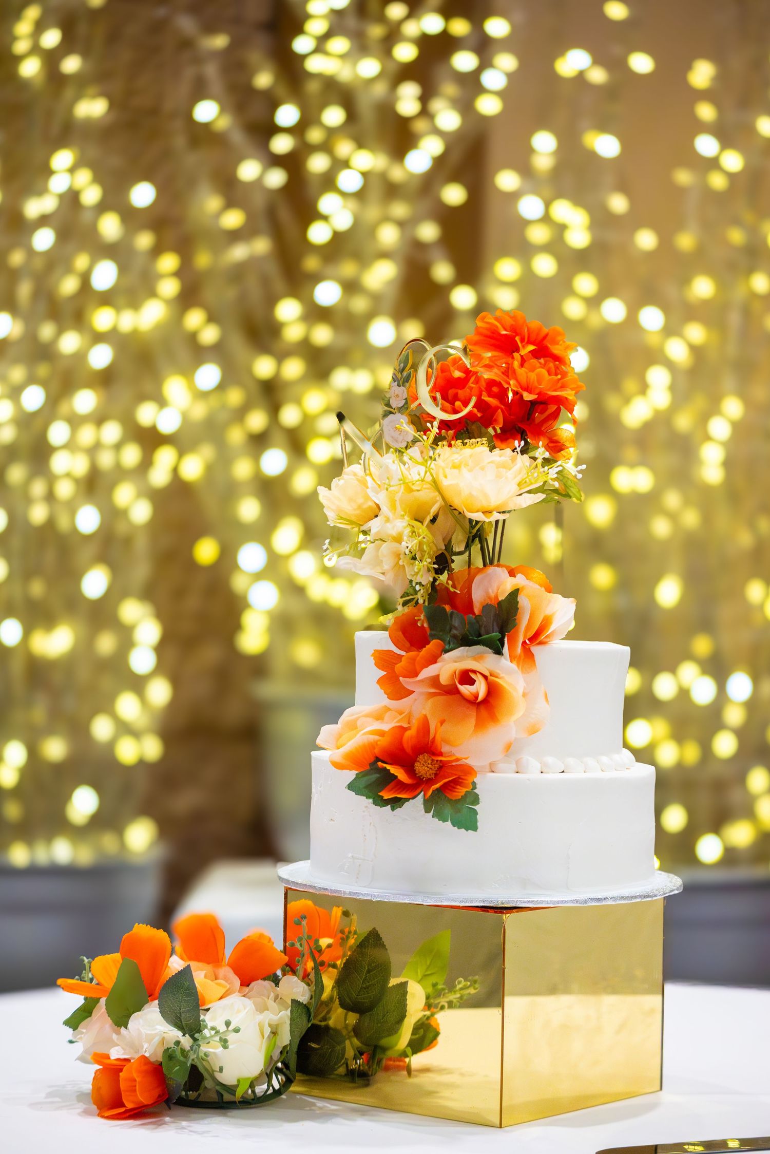 Three-tier white wedding cake adorned with orange and cream flowers on gold stand, bokeh light backdrop.
