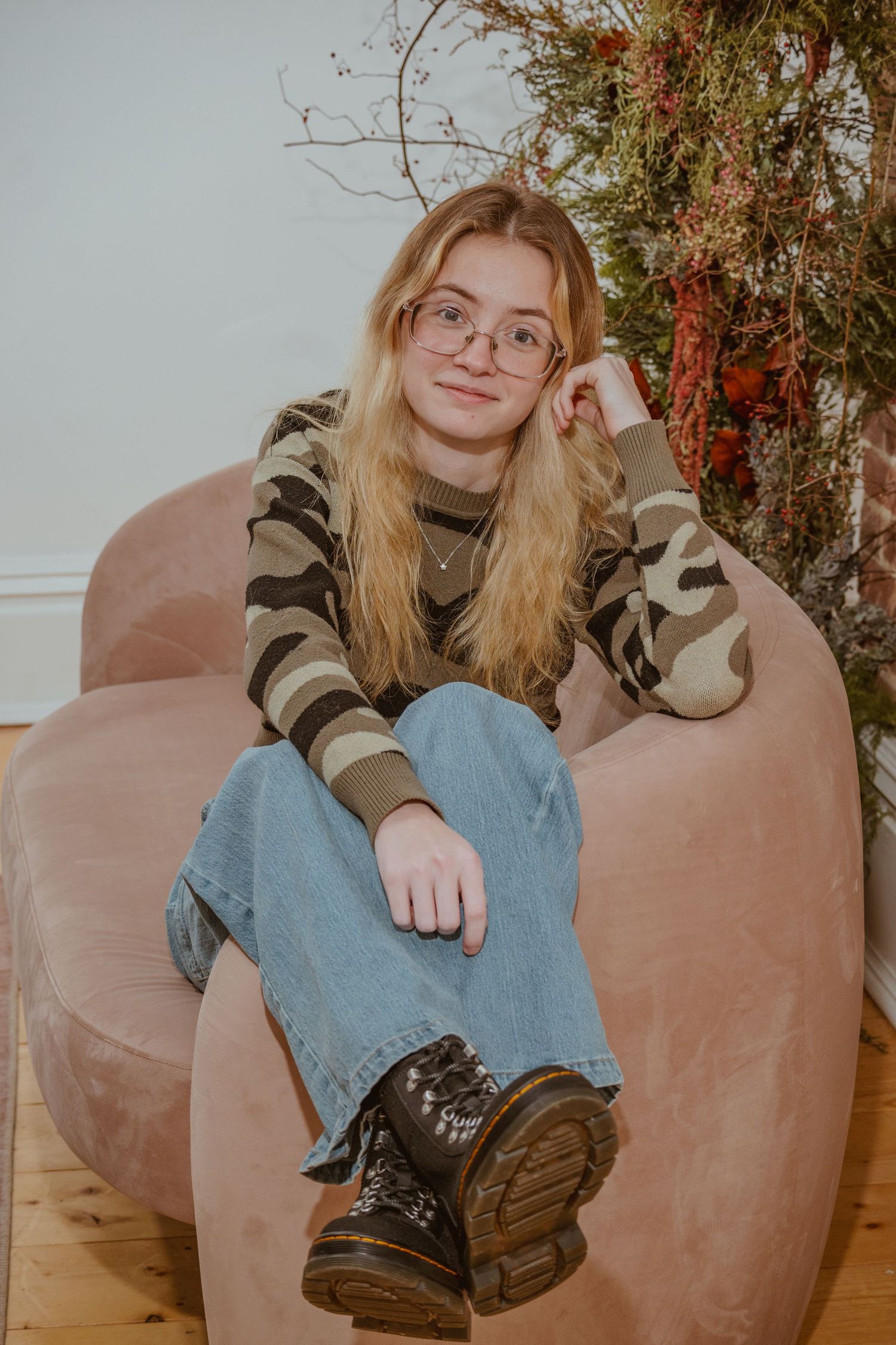 Young woman with blonde hair and glasses sits relaxed in a pink chair wearing camo top, jeans, and Dr. Martens boots.
