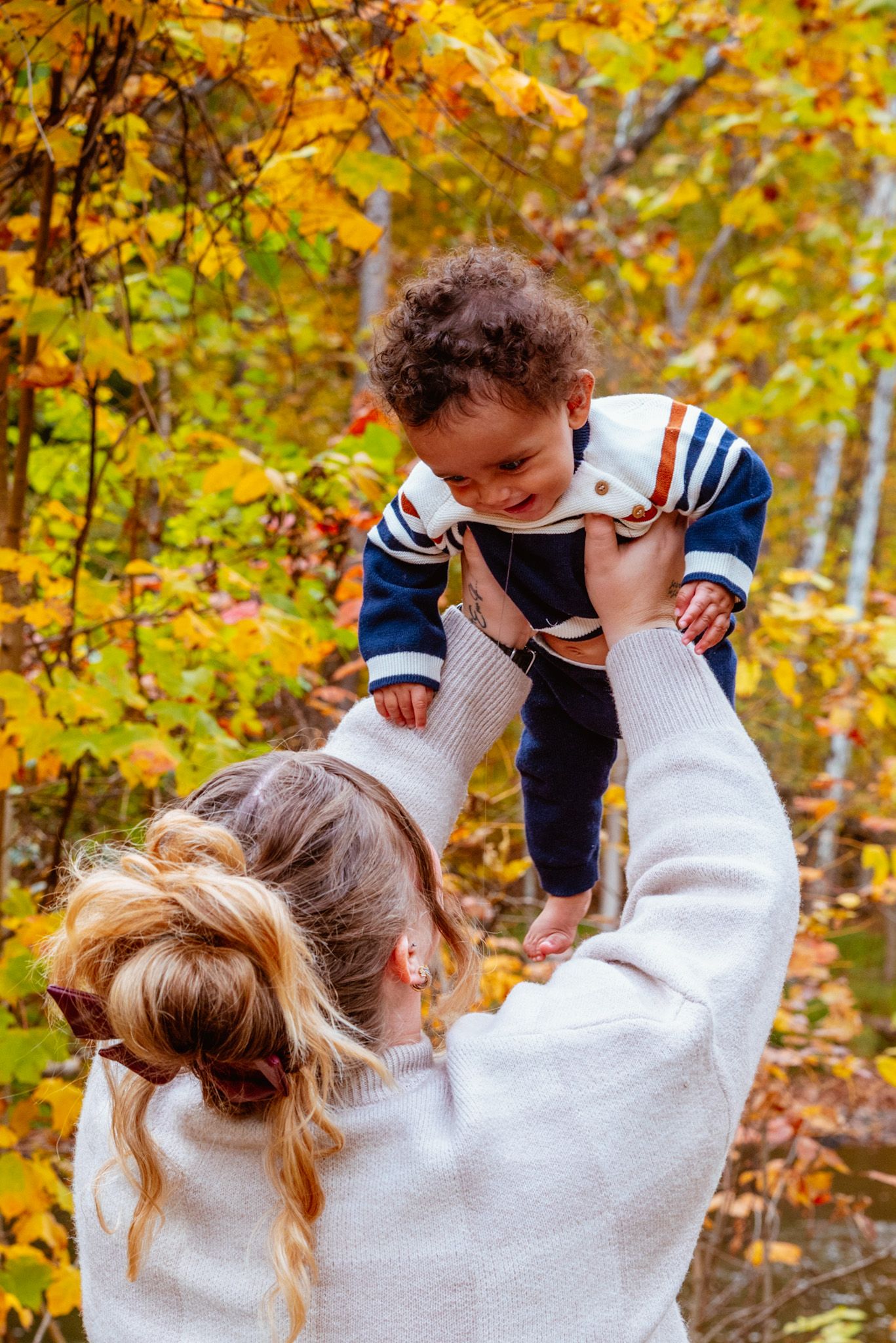 Mother lifting smiling baby in autumn forest with colorful yellow and orange fall foliage in background.