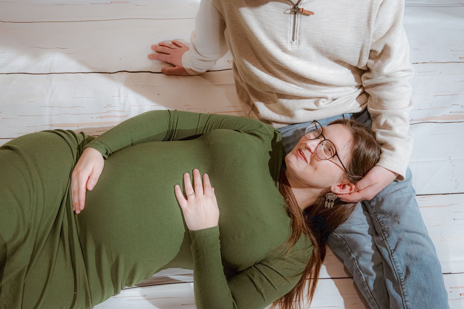 Pregnant woman lying on floor while partner in white sweater gently touches her baby bump during maternity photoshoot.