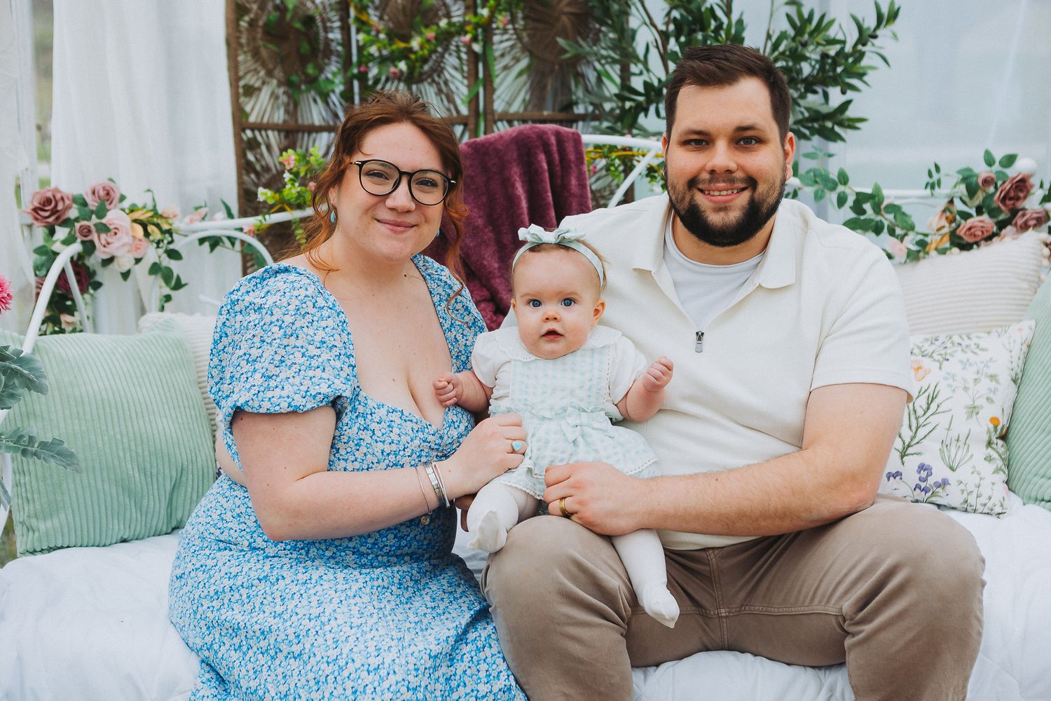 Family sits together on outdoor patio furniture with floral pillows and greenery in background.