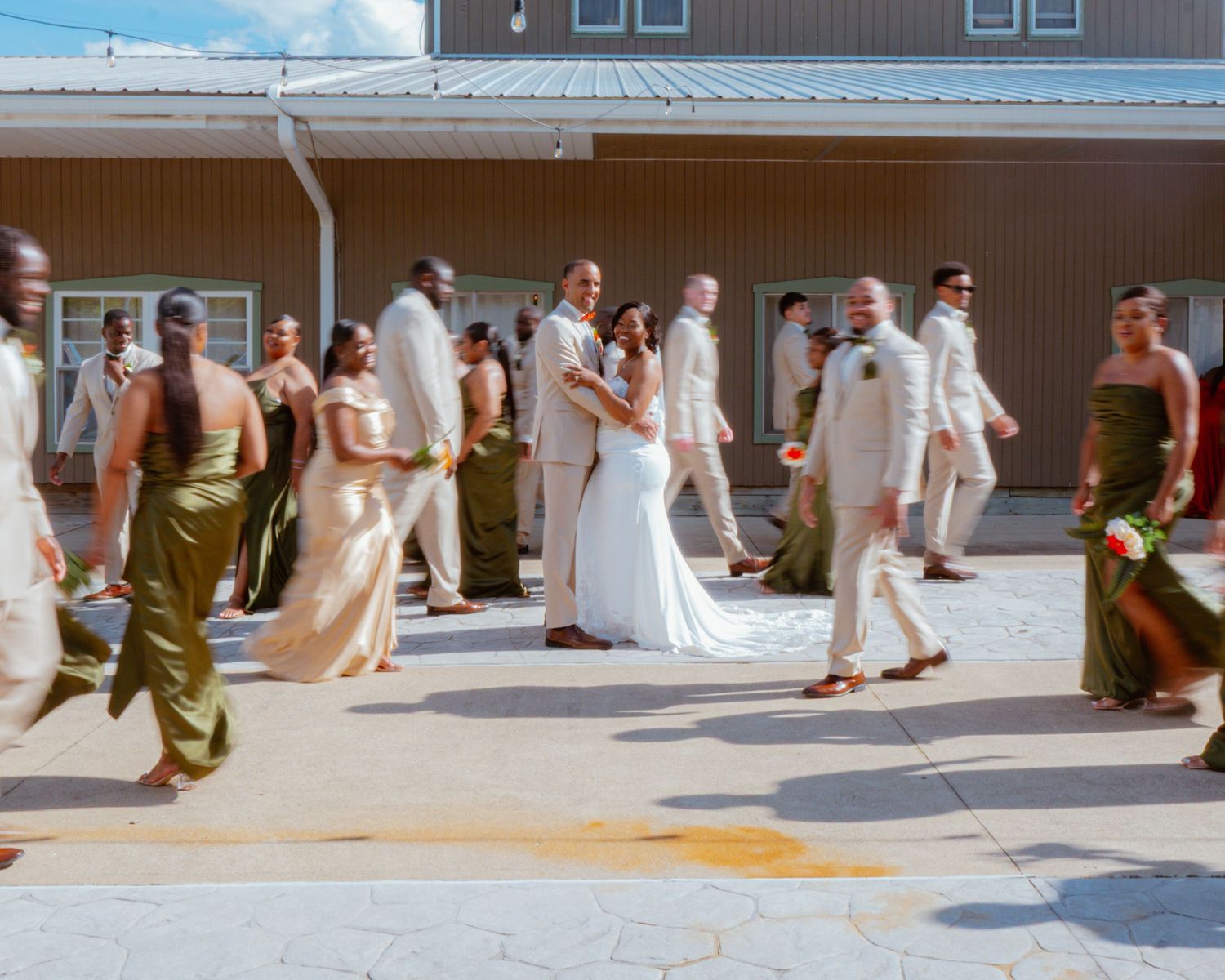 Wedding party in olive green and beige attire walking together outside a brown building on a sunny day.