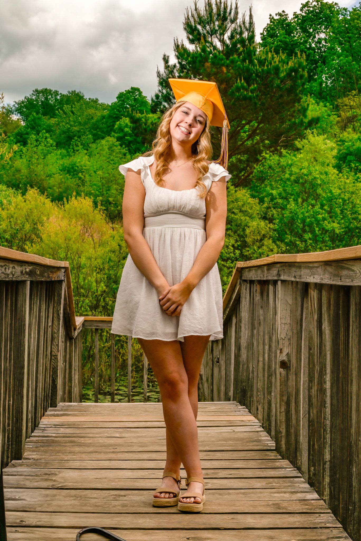Smiling graduate in white dress and gold cap poses on wooden bridge surrounded by lush green trees.