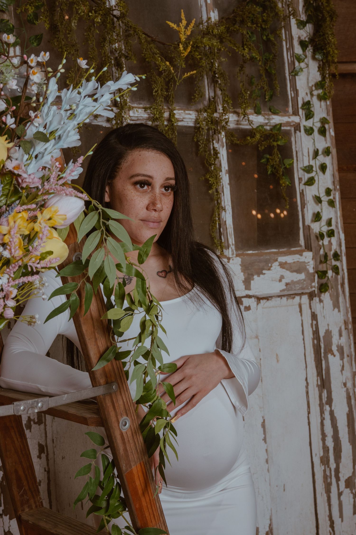 Pregnant woman in white dress poses by a rustic door adorned with lush greenery and spring flowers.