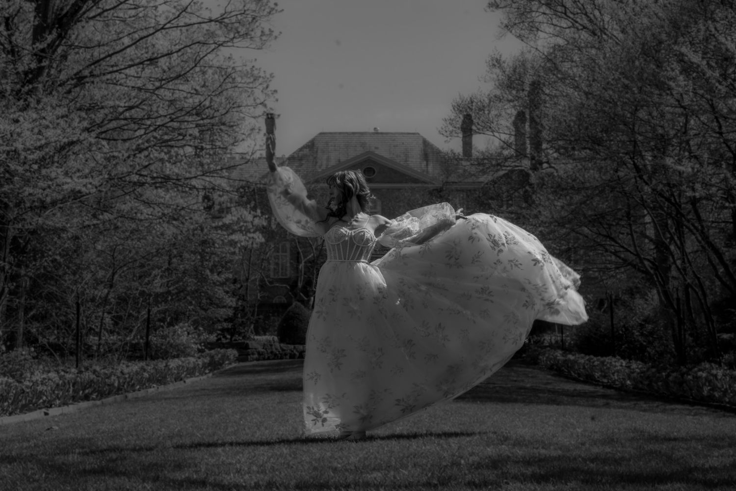 Black and white photograph showing a figure in a flowing white dress leaping gracefully in mid-air on a wooded path.