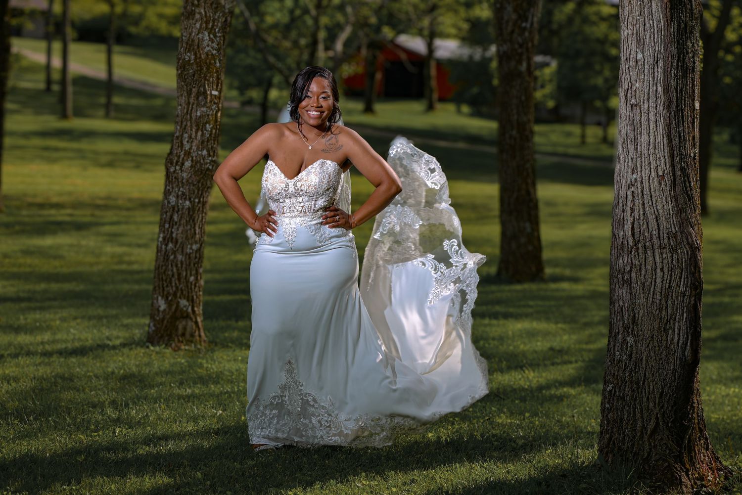 Bride in elegant white lace mermaid wedding gown with veil posing outdoors between trees on green lawn.