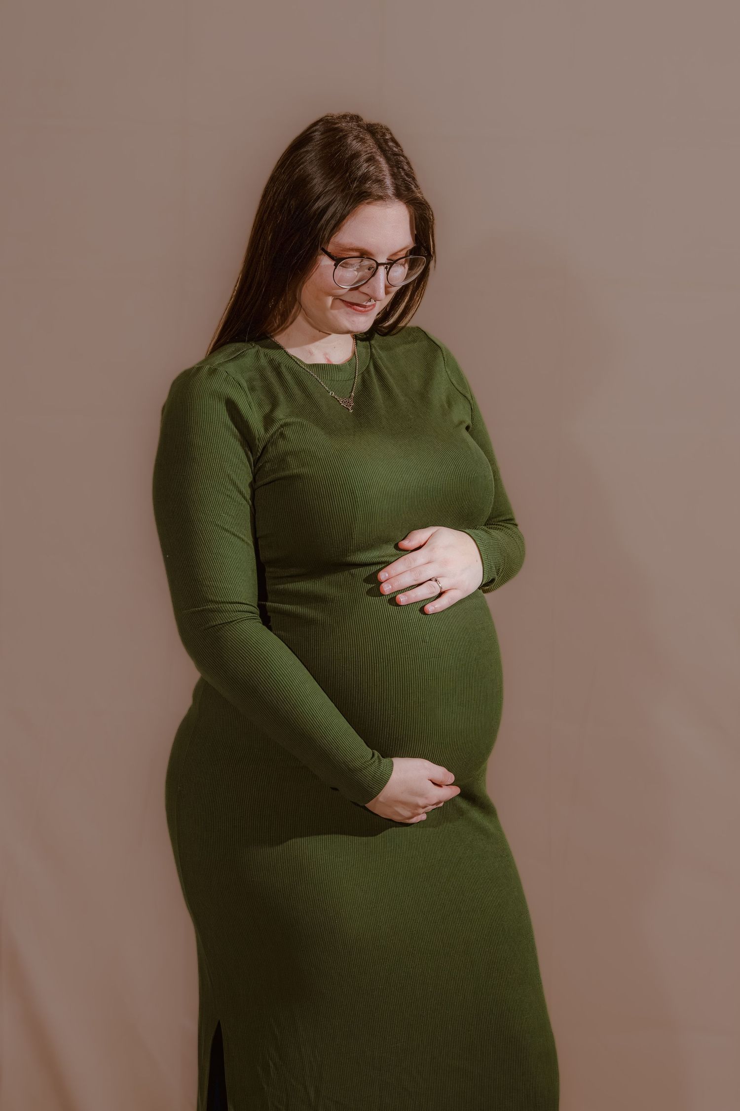 Pregnant woman in green ribbed dress cradling baby bump, wearing glasses, studio portrait on beige background.