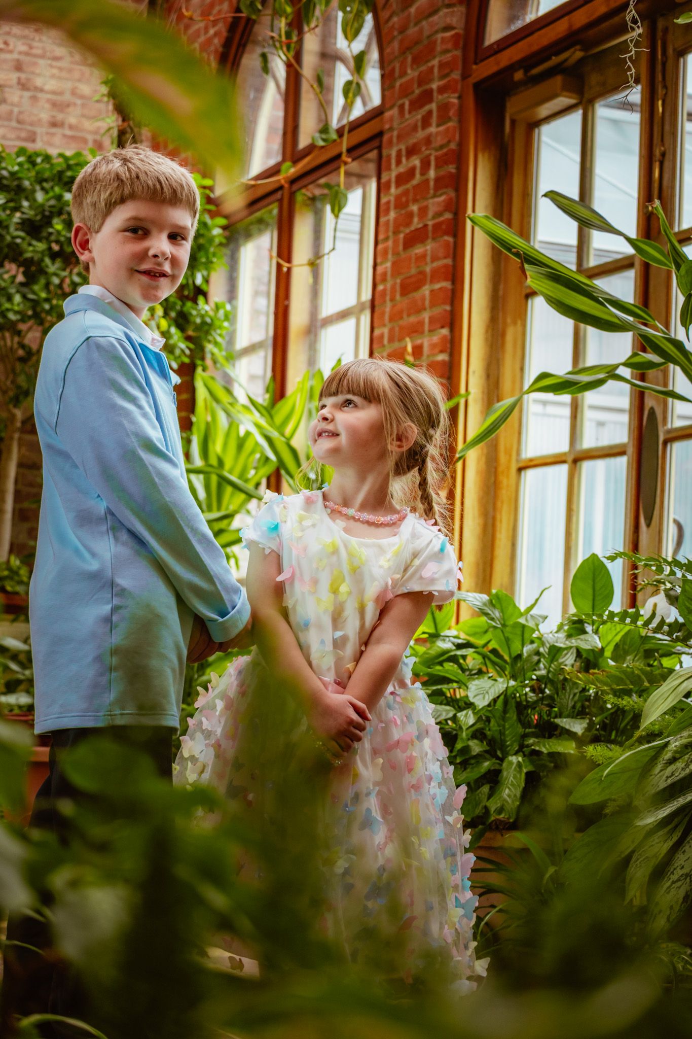 Two children in formal attire stand together in a greenhouse setting with lush plants and brick walls.
