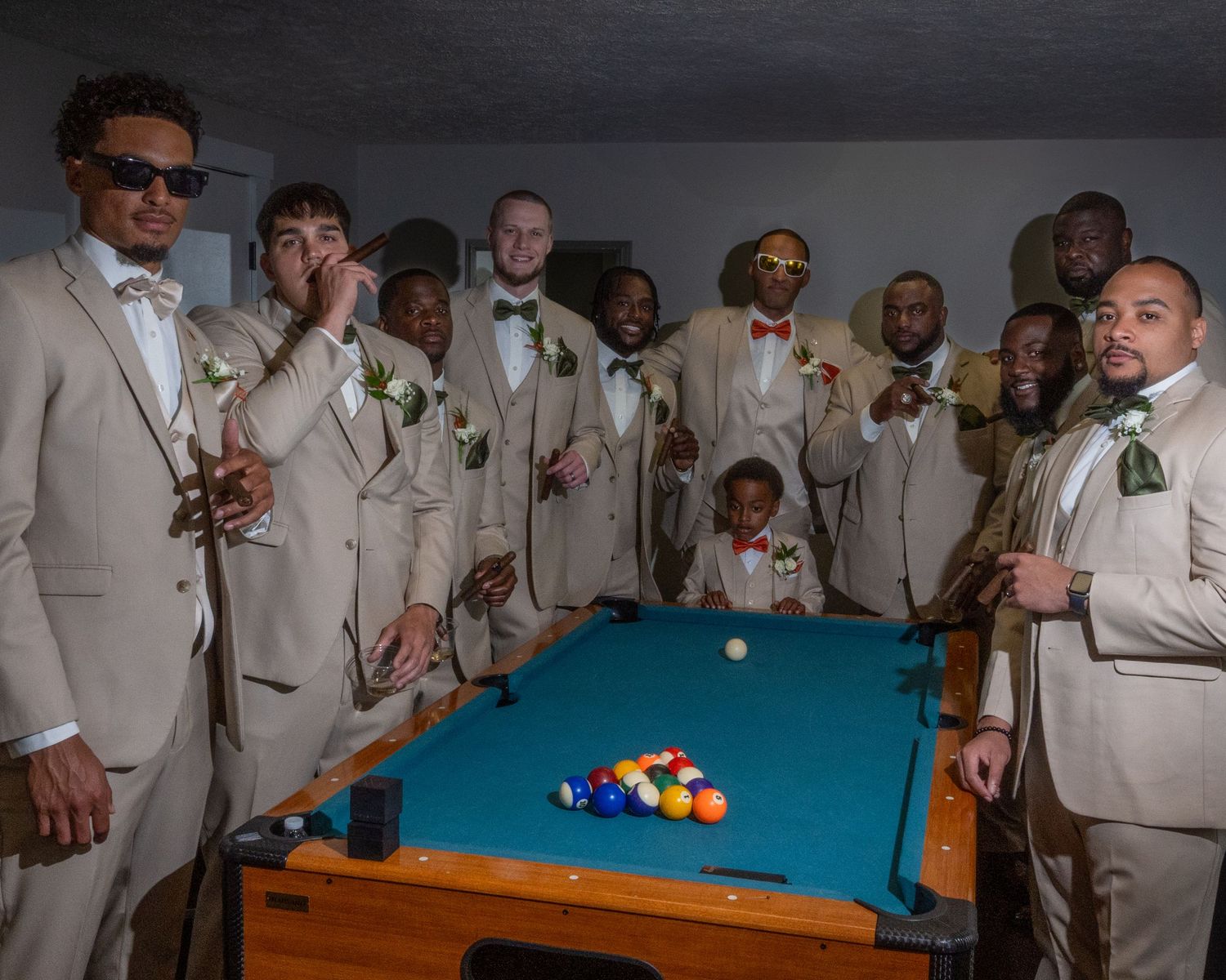 Groomsmen in tan suits with boutonnieres pose around a pool table with colorful billiard balls at a wedding.