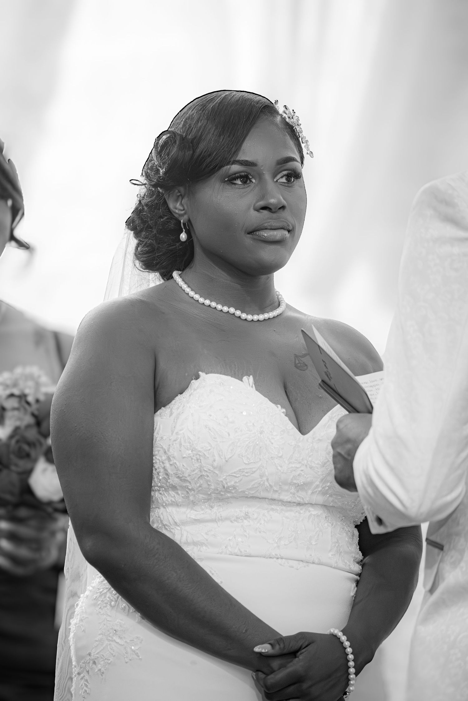 Black and white photo of a bride in a strapless lace gown wearing pearl jewelry and a tiara at her wedding ceremony.