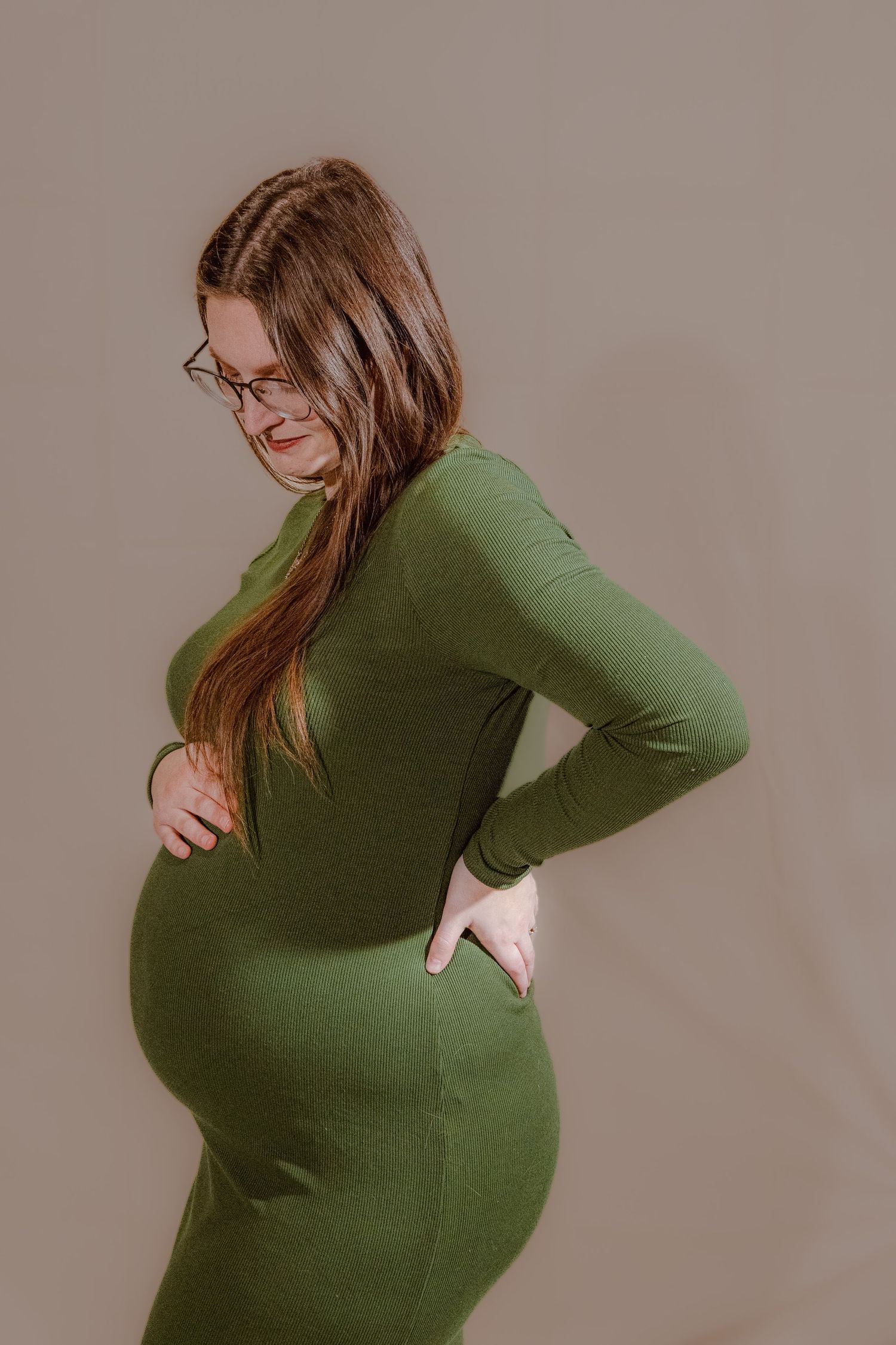 Pregnant woman in green long-sleeve dress holding belly, wearing glasses, posing against neutral background.