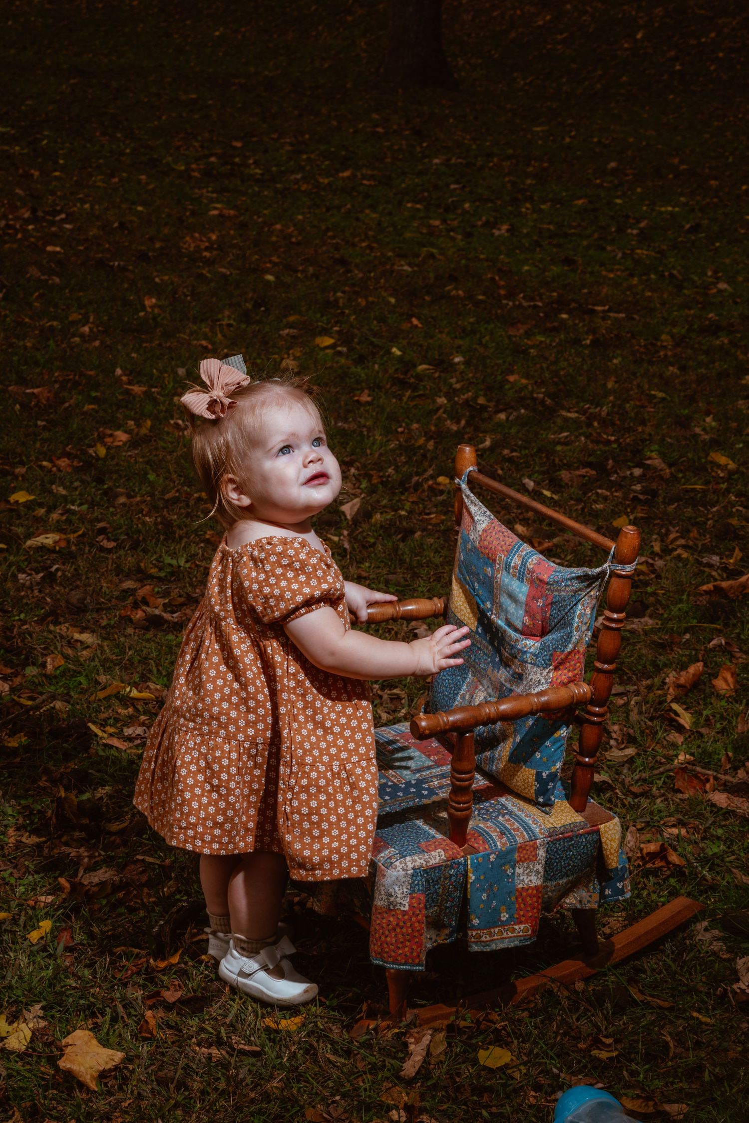Toddler girl in polka dot dress standing beside a wooden rocking chair with quilt in autumn park.