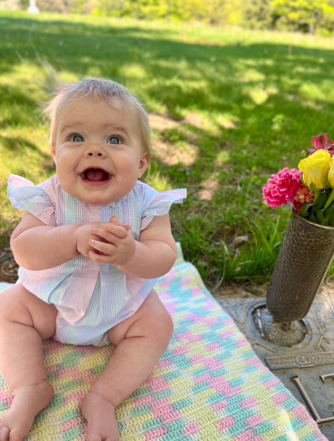A joyful baby sits on a colorful crocheted blanket outdoors on grass, wearing a light-colored dress and bow.