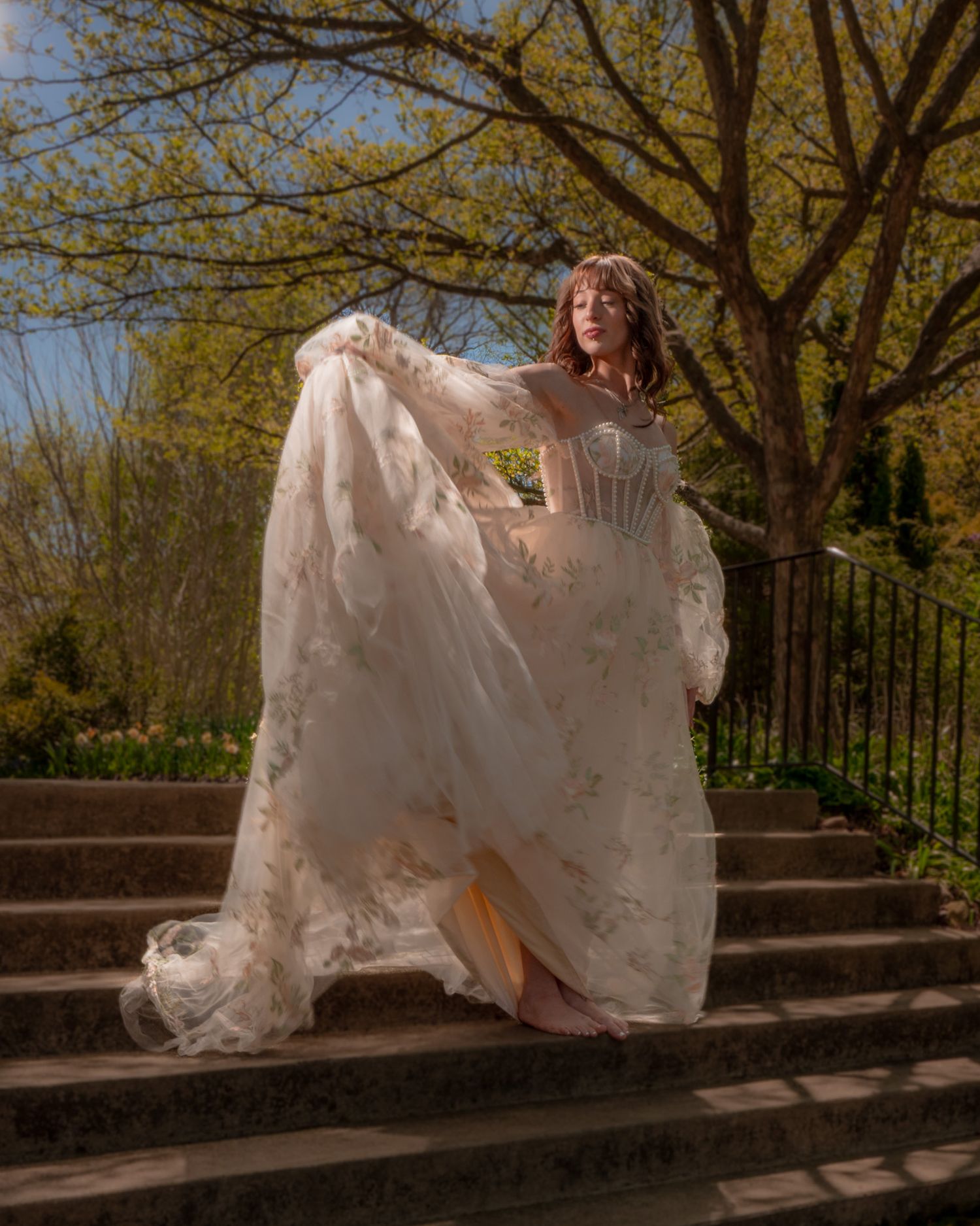 Woman in flowing lace bridal gown poses gracefully on stone steps surrounded by spring trees.