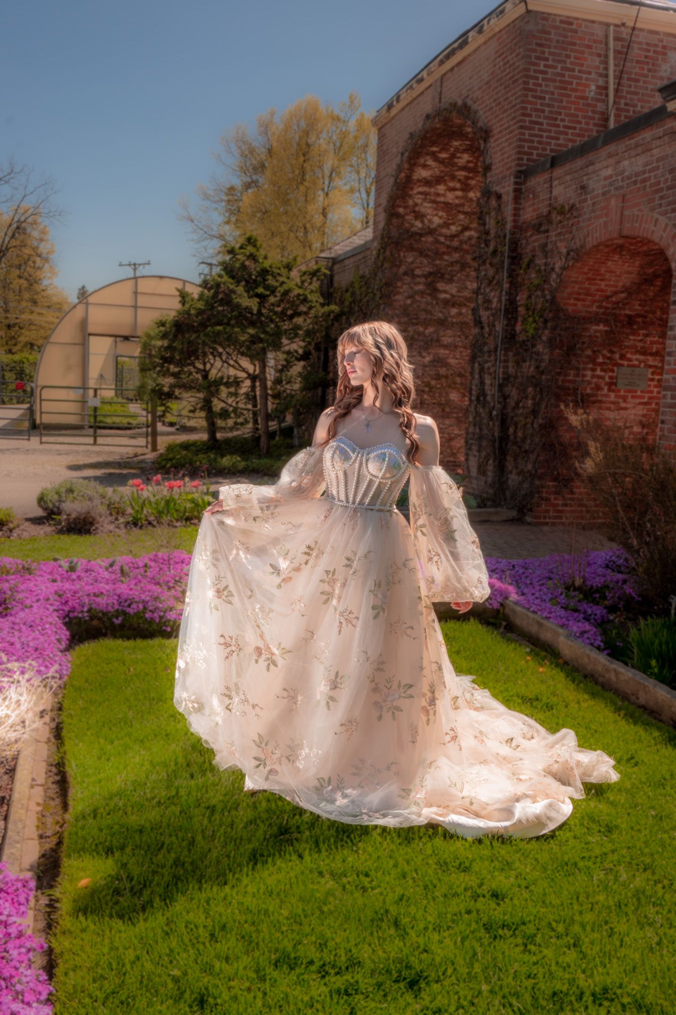 A romantic vintage-style wedding dress with lace details flows gracefully in a garden setting with purple flowers and brick walls.