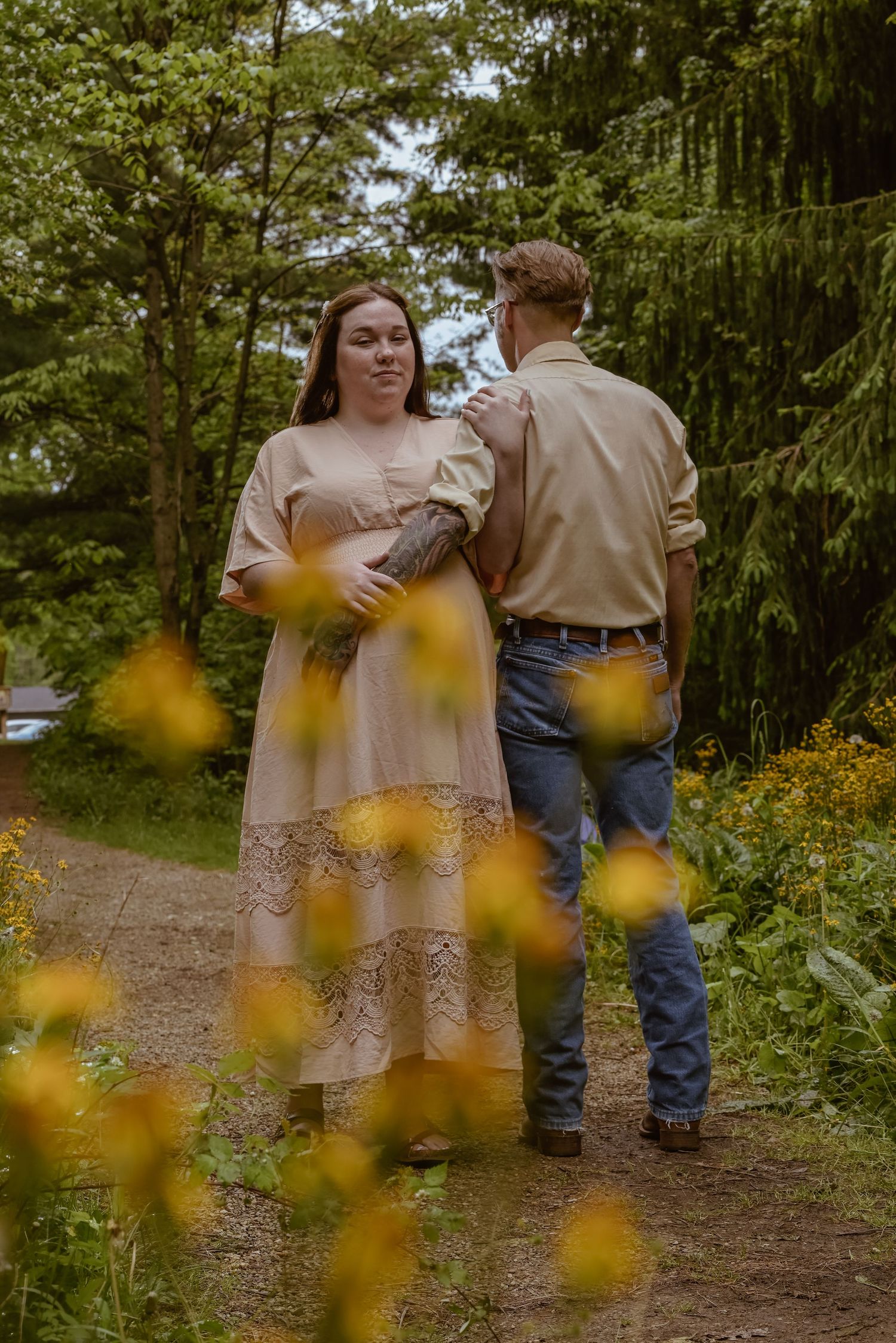 Couple walking on a forest path, woman in beige lace dress holding flowers, man in tan shirt and jeans, yellow wildflowers foreground.