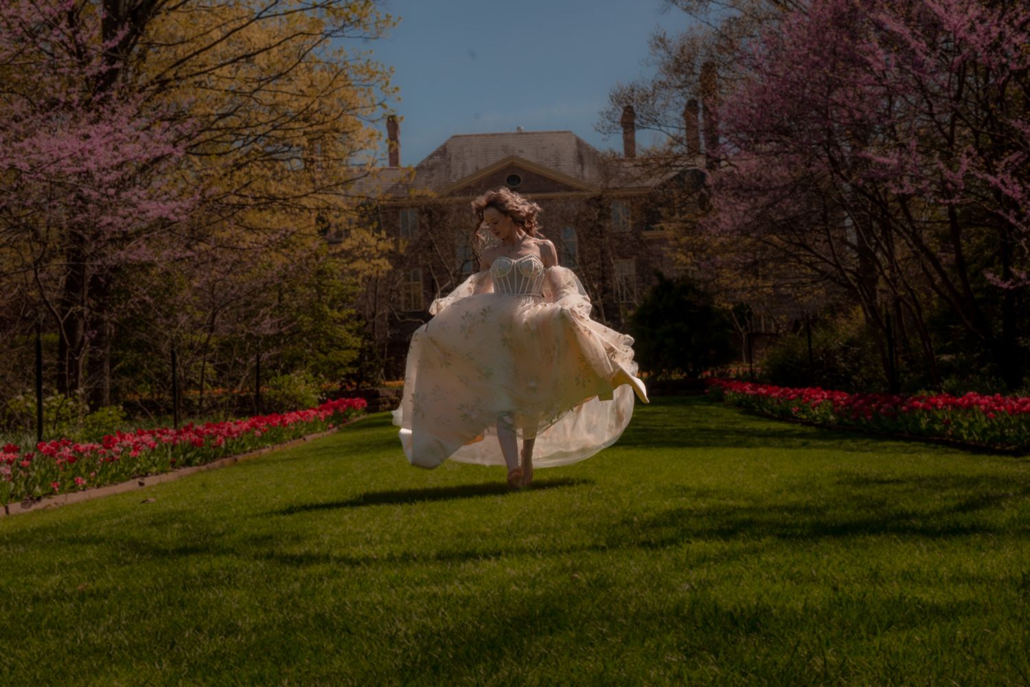 Bride twirling in a flowing white gown on green grass surrounded by blooming spring trees and tulips outside an estate.
