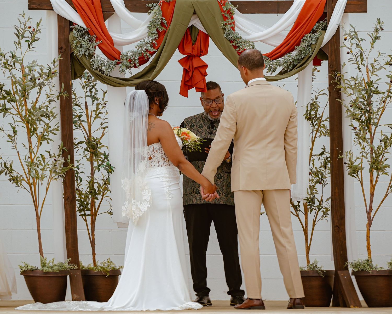Bride and groom holding hands at outdoor wedding ceremony beneath floral arch with greenery and red fabric draping.
