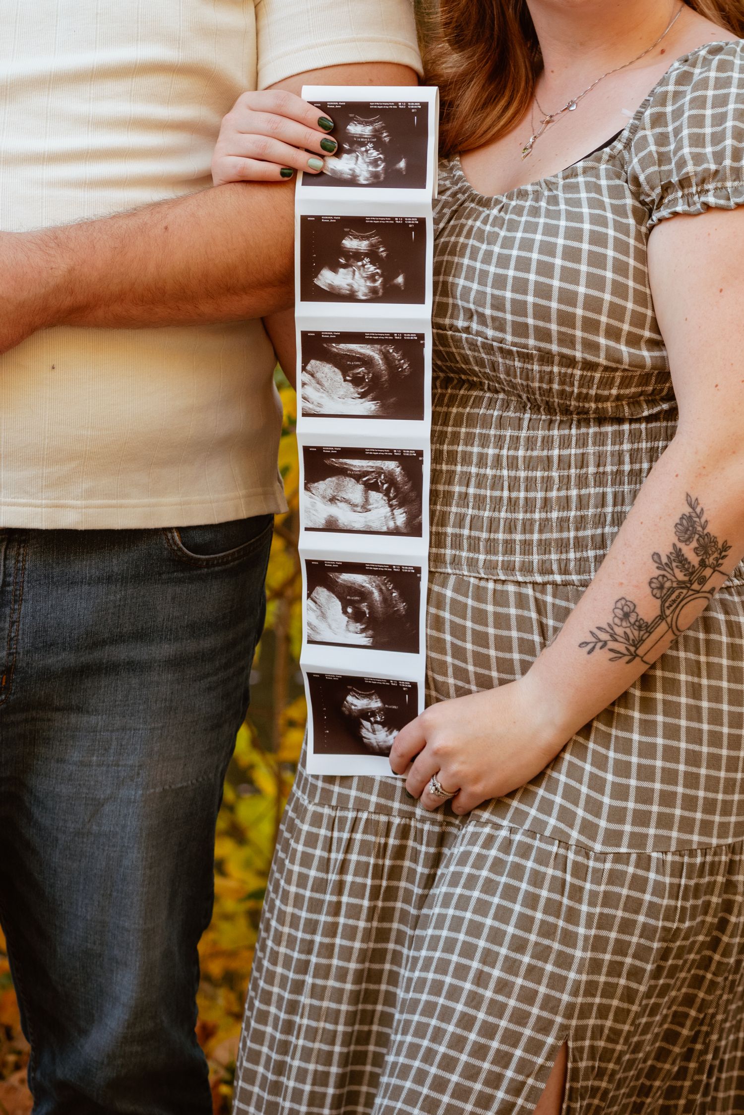 Couple holding ultrasound strip photos outdoors, woman in plaid dress with floral tattoo announcing pregnancy.