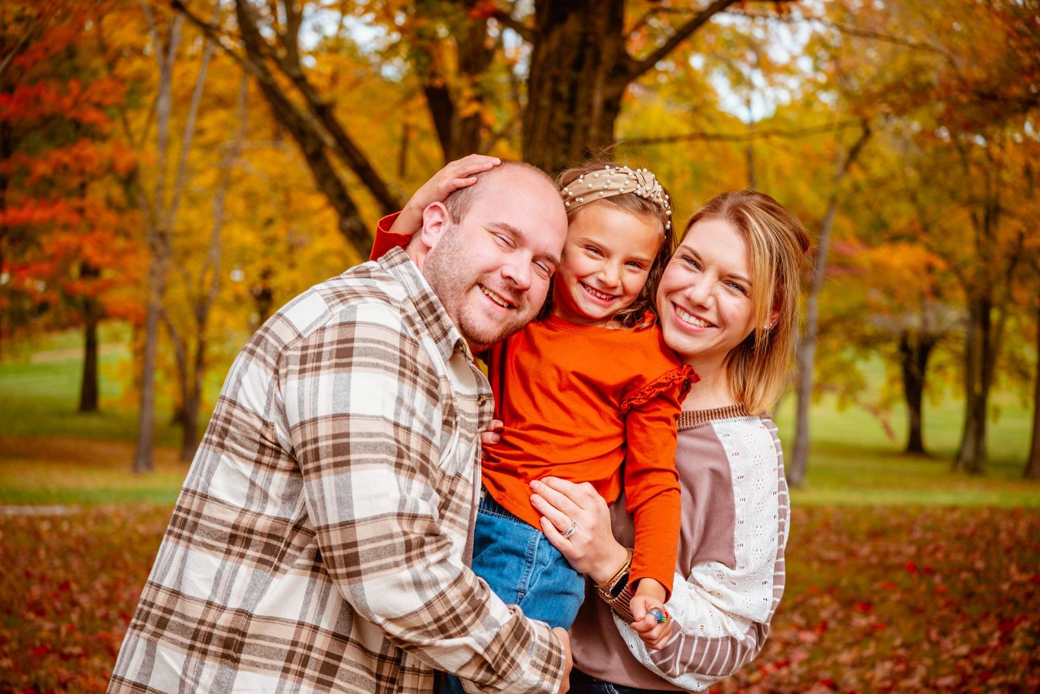 Happy family of three posing outdoors in autumn park with colorful fall foliage in background.