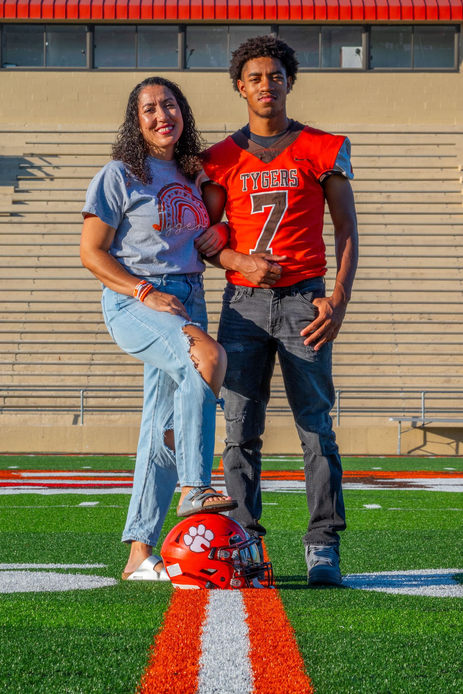 A proud mom poses with her son wearing a Tigers #7 football jersey on a stadium field with a helmet.