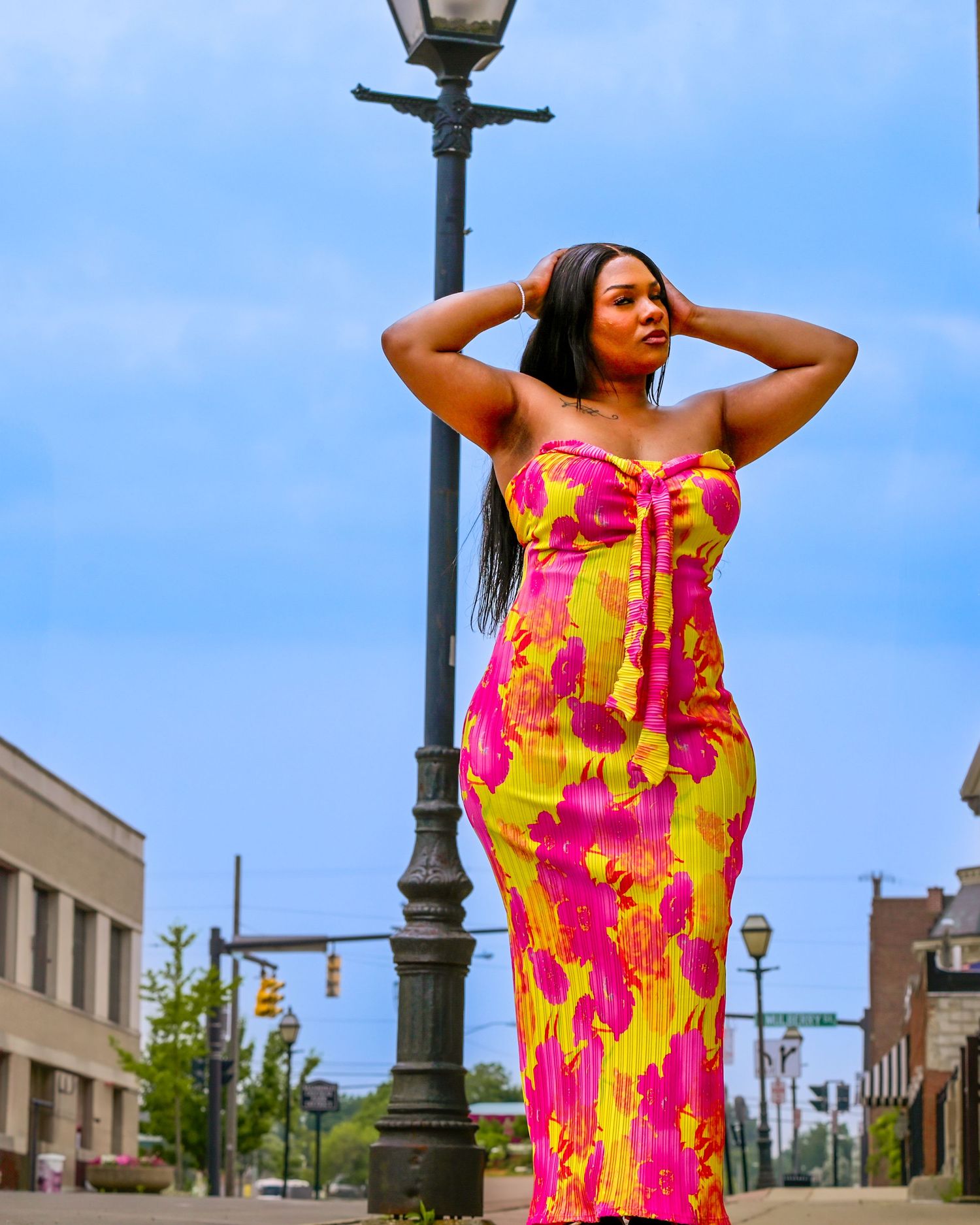 A model poses in a vibrant pink and yellow floral print dress against a blue sky on a city street.