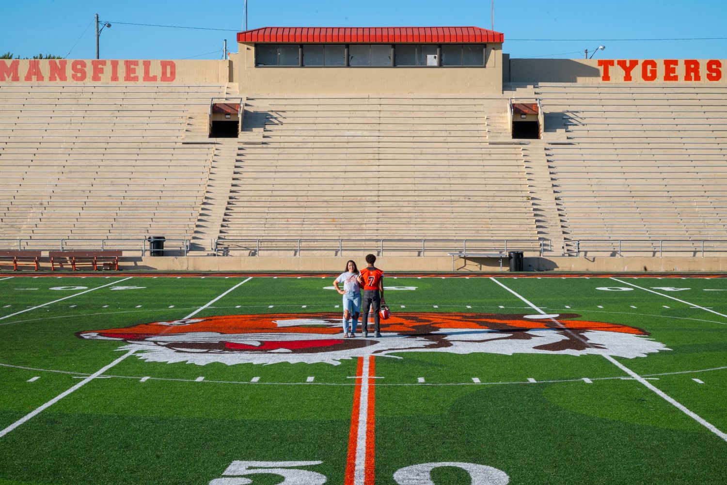 Couple standing on the 50-yard line of Mansfield Tygers football stadium with empty bleachers behind them.