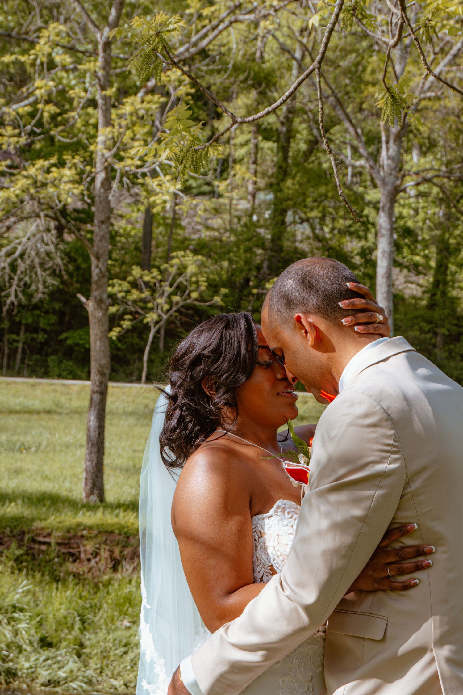A bride and groom share a tender kiss outdoors surrounded by lush green trees on their wedding day.