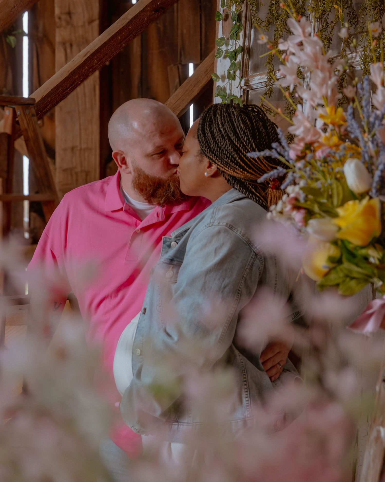 Couple sharing a tender kiss in a rustic barn surrounded by colorful spring flowers, man in pink shirt, woman in denim jacket.