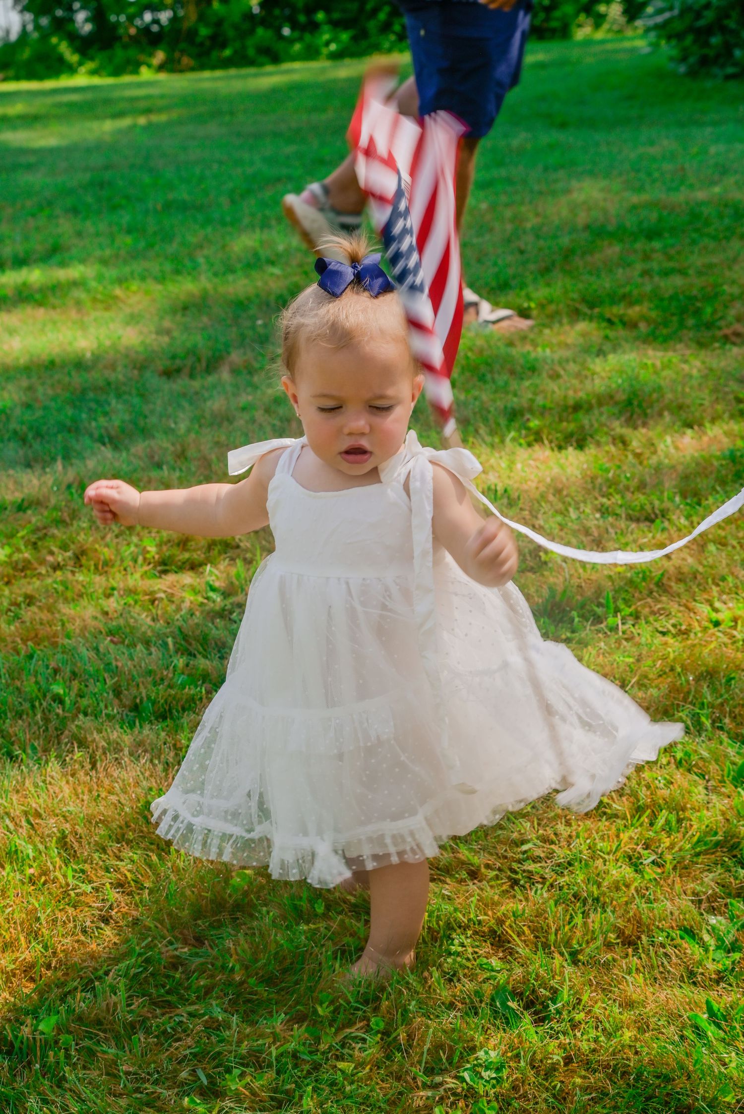 Toddler girl in white dress walking barefoot on green grass with American flag in background.