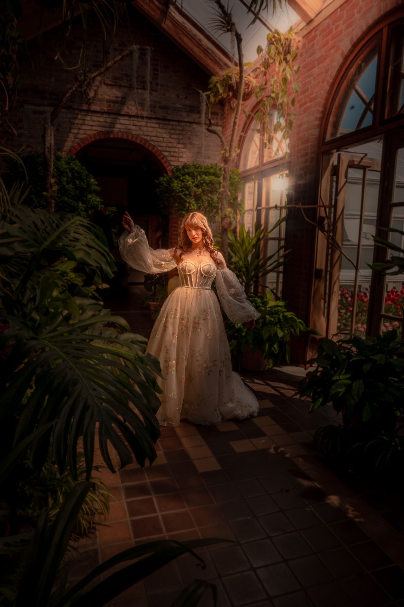 Woman in white gown standing in sunlit greenhouse with lush tropical plants and brick arched windows.