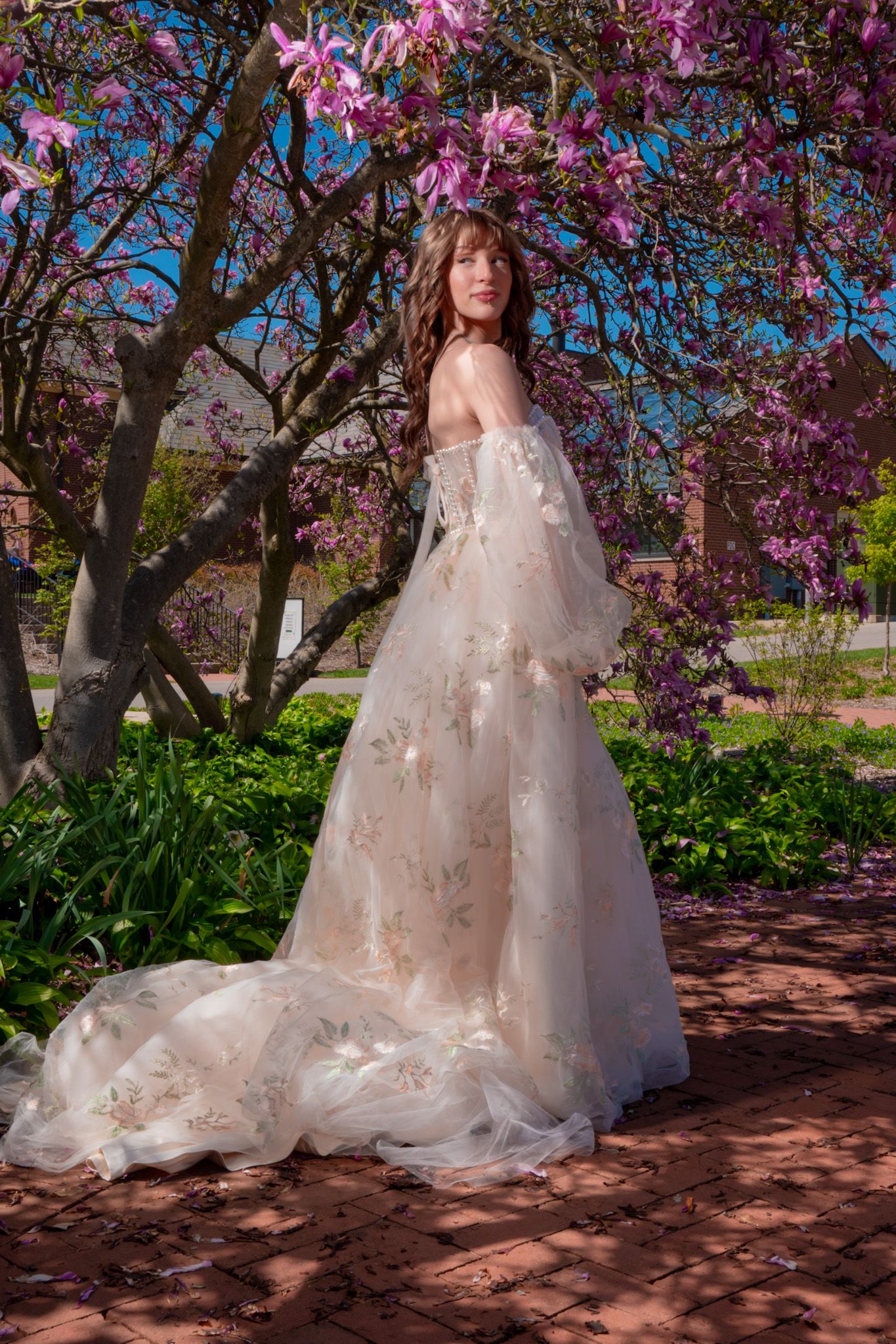 An elegant off-shoulder lace wedding dress with flowing train photographed under blooming pink magnolia trees.